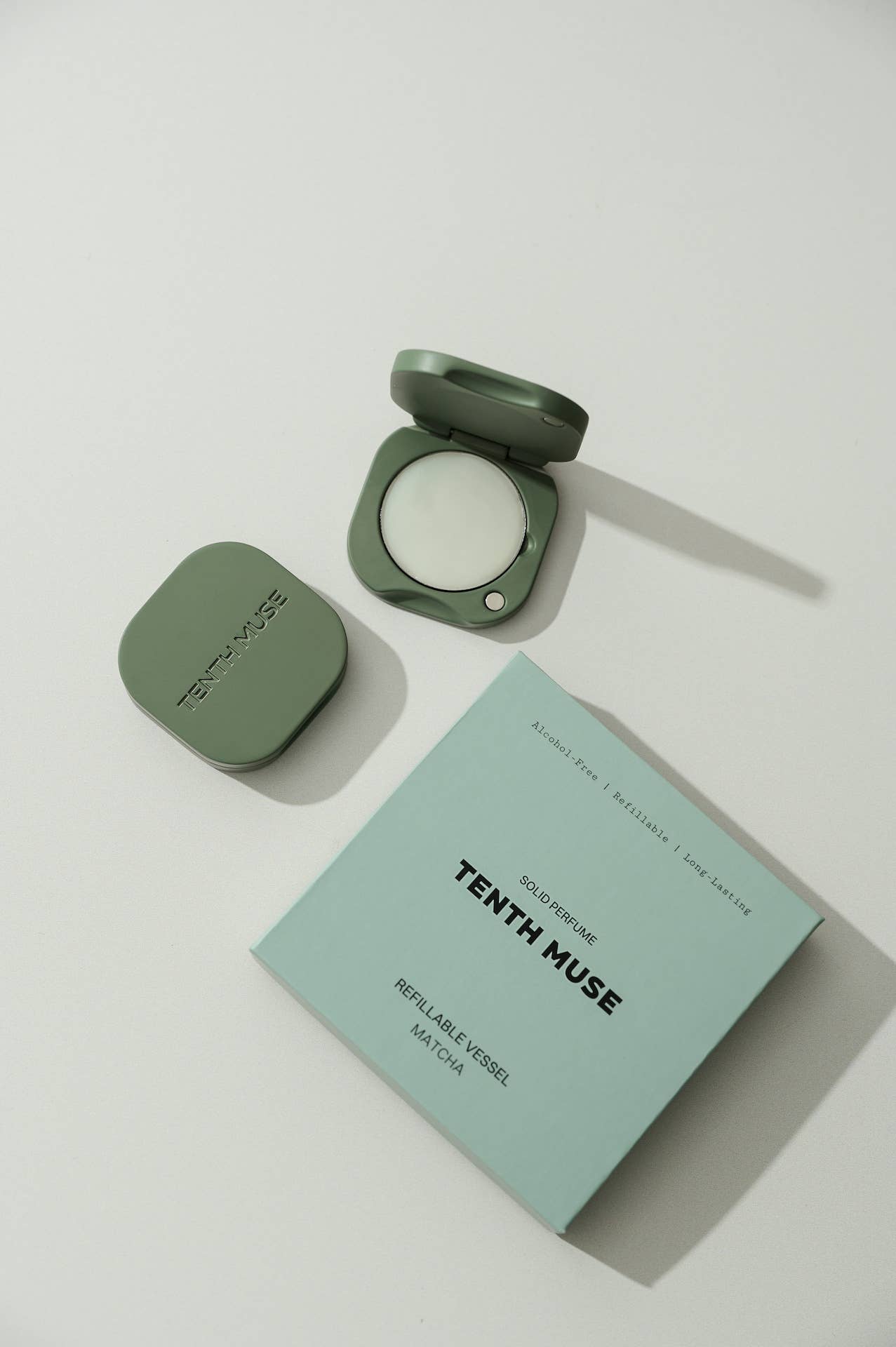 A green/matcha square refillable compact for the tenth muse solid perfume balms that has 'TENTH MUSE' lightly carved into the compact sits next to an open compact that has a balm inside of it, beside a white square packaging box for the Tenth Muse refillable vessel. At the top of the box, all in black, it says Alcohol-free, Refillable, Long-lasting'. Beneath that it says Solid Perfume and underneath  'TENTH MUSE'. At the bottom of the box it says Refillable Vessel and Matcha beneath, that being the shade. 