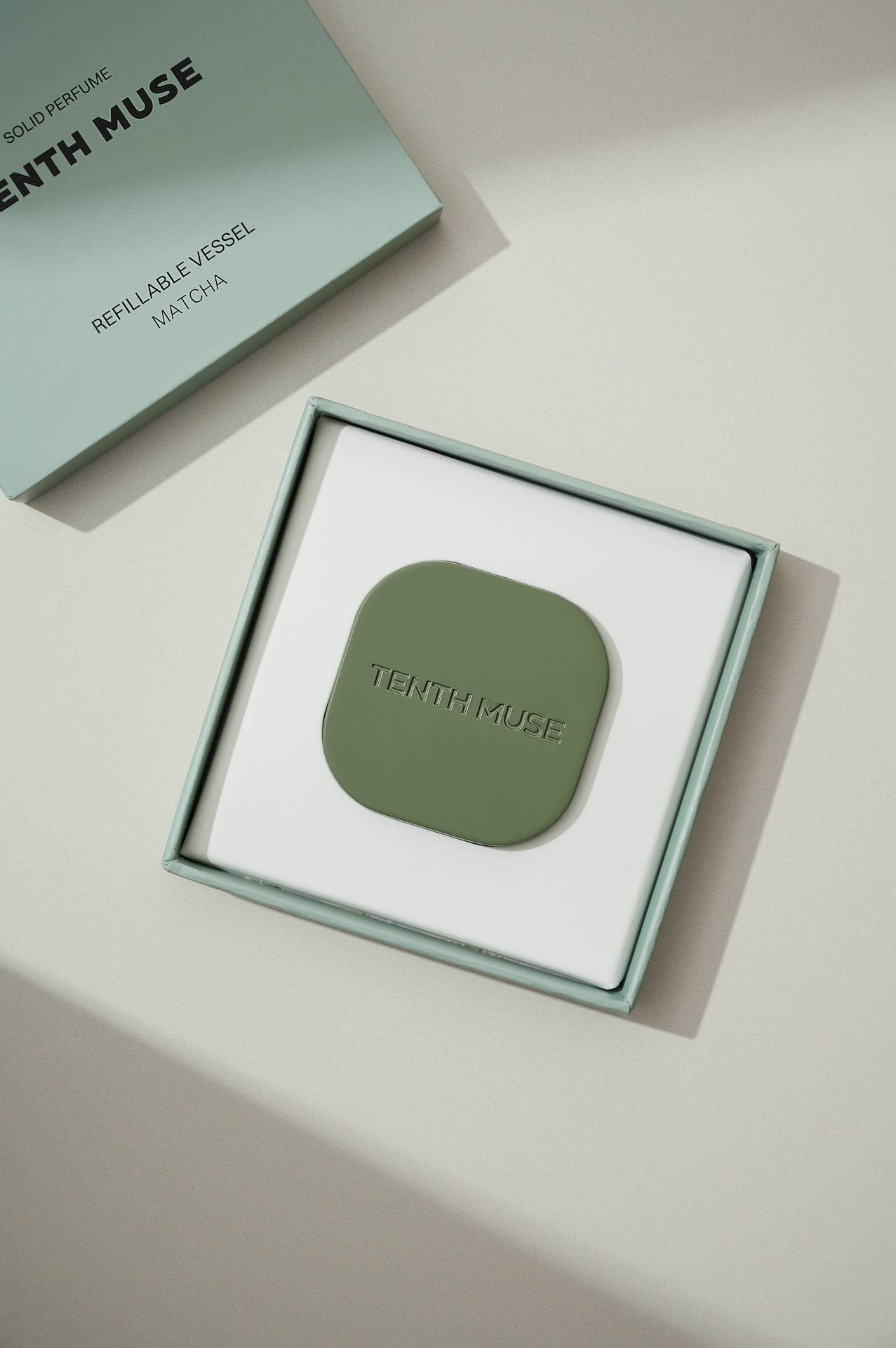 A green/matcha square refillable compact for the tenth muse solid perfume balms. The compact is sitting inside of the packaging that the compact comes in which is a square box that is white on the inside and has 'TENTH MUSE' lightly carved into the compact. Next to the open box is the lid of the compact box which is also green/matcha and says TENTH MUSE in black followed by Refillable Vessle and underneath that Matcha, the shade of the compact.