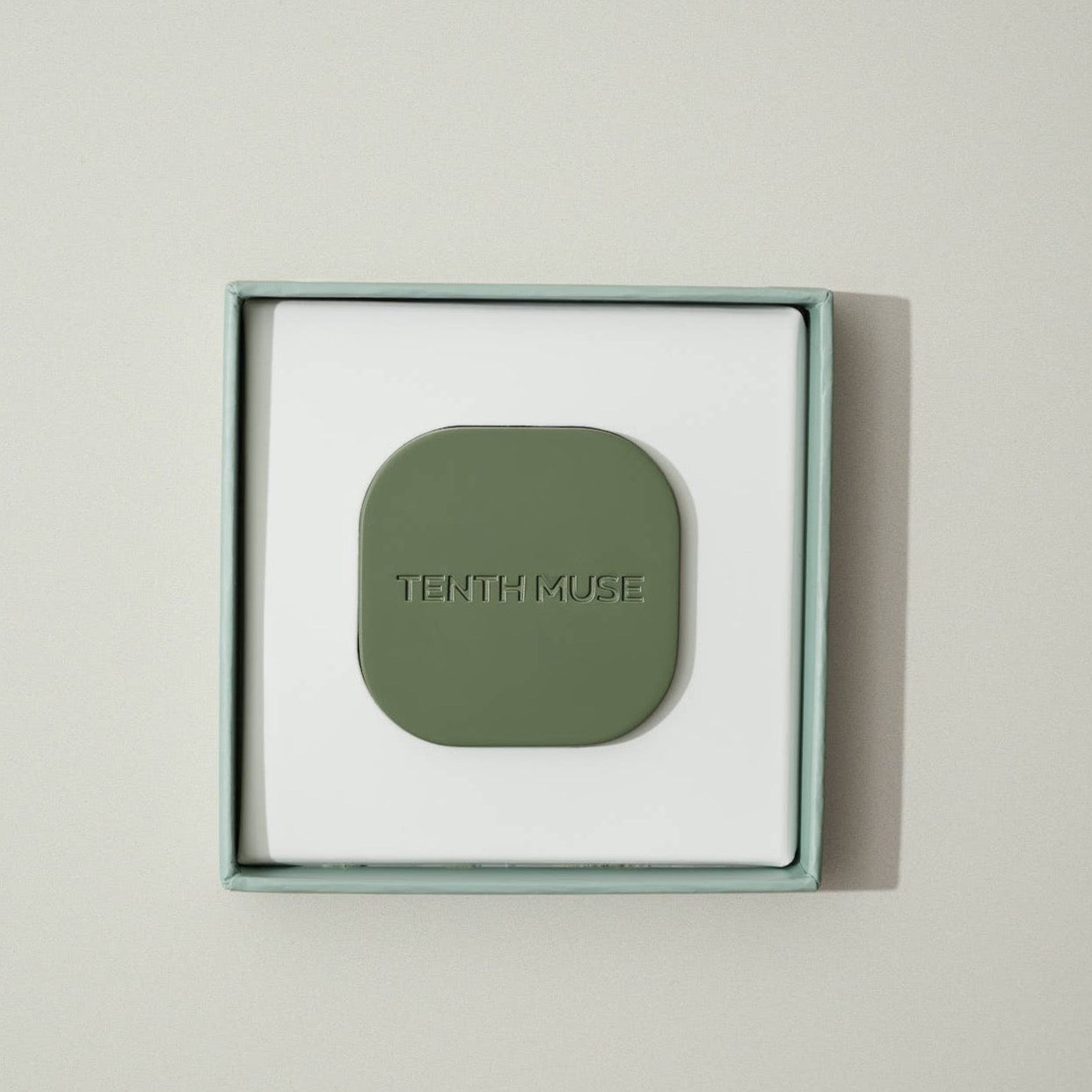 A green/matcha square refillable compact for the tenth muse solid perfume balms. The compact is sitting inside of the packaging that the compact comes in which is a square box that is white on the inside with a green/matcha and has 'TENTH MUSE' lightly carved into the compact.