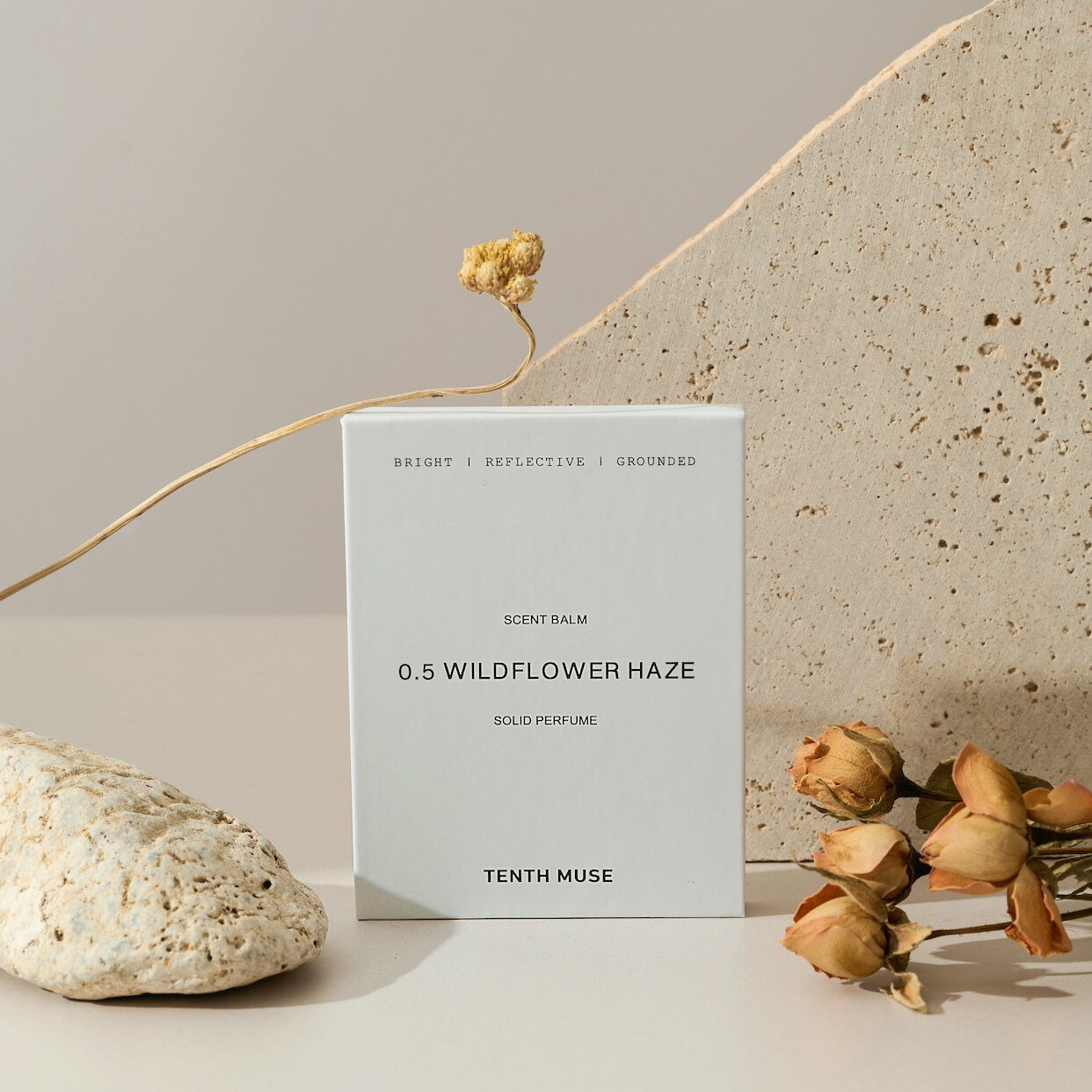 A white square packaging box for the Tenth Muse solild perfume balm sits next to a clay rock and some dried peach flowers. The box is white and at the top in black block letters says 'bright, reflective, grounded', describing the scent. Beneath that it says Refill and beneath that '0.5 WILDFLOWER HAZE', which is the scent. Underneath that it says 'sold perfume balm' followed by TENTH MUSE at the bottom of the packaging.