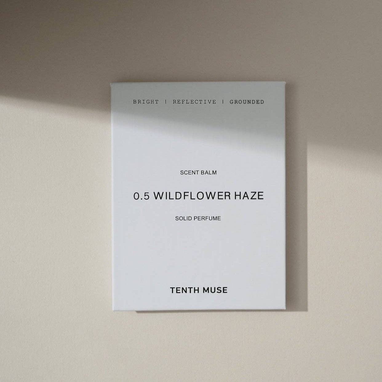 A white square packaging box for the Tenth Muse solild perfume balm. The box is white and at the top in black block letters says 'bright, reflective, grounded', describing the scent. Beneath that it says Refill and beneath that '0.5 WILDFLOWER HAZE', which is the scent. Underneath that it says 'sold perfume balm' followed by TENTH MUSE at the bottom of the packaging.