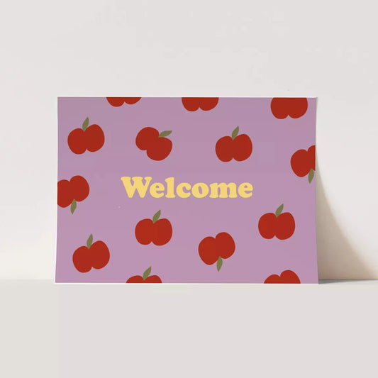 An A5 postcard with a pastel purple background featuring a scattered pattern of red apples with green leaves. The word "Welcome" is printed in bold, yellow letters in the center.