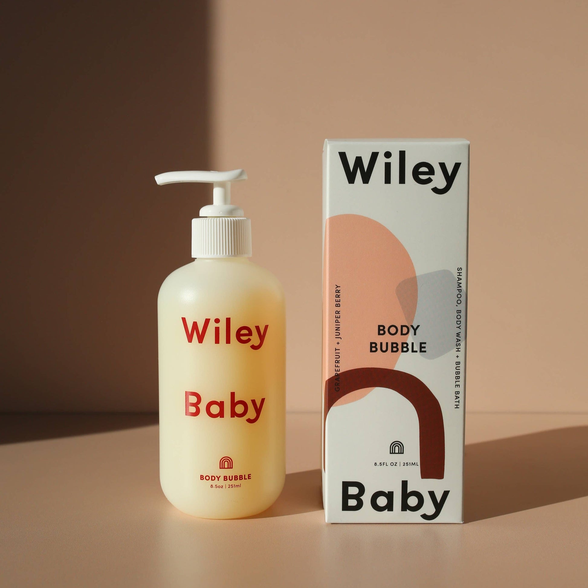 A clear bottle with a white refillable bump with the Wiley Baby Body Bubble inside, making the bottle appear yellow. In red letters it says Wiley Baby Body Bubble. Next to the bottle is the packaging which is white with pink, blue and red abstract shapes. At the top it says Wiley in black and at the bottom it says Baby. Along the side of the bottle, horizontally, it says Grapefruit + Juniper Berry and on the other side it says Shampoo, Body Wash + Bubble Bath. This is against a pink background. 