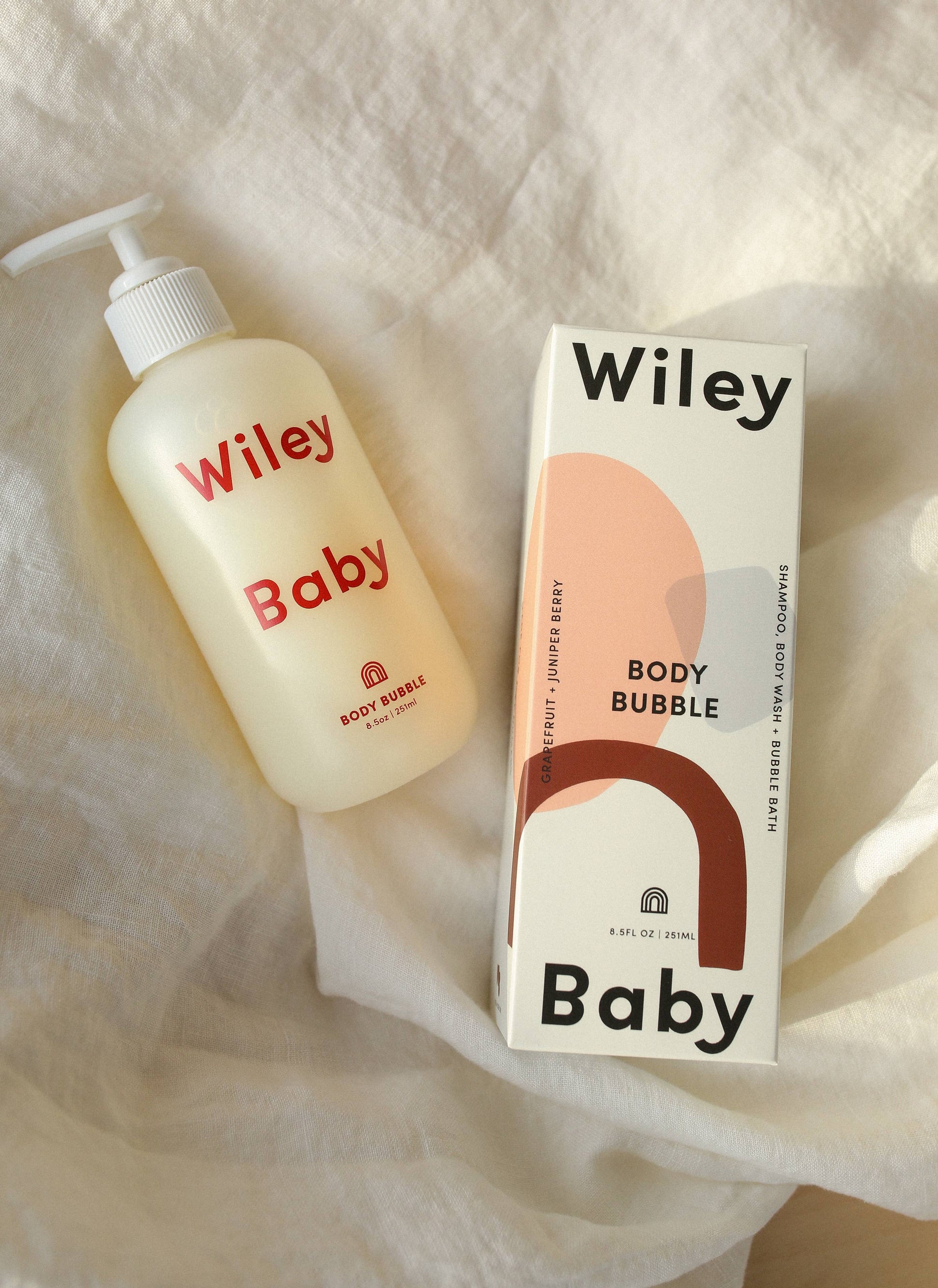 A clear bottle with a white refillable bump with the Wiley Baby Body Bubble inside, making the bottle appear yellow lays on top of white sheets. In red letters it says Wiley Baby Body Bubble. Next to the bottle is the packaging which is white with pink, blue and red abstract shapes. At the top it says Wiley in black and at the bottom it says Baby. Along the side of the bottle, horizontally, it says Grapefruit + Juniper Berry and on the other side it says Shampoo, Body Wash + Bubble Bath. 