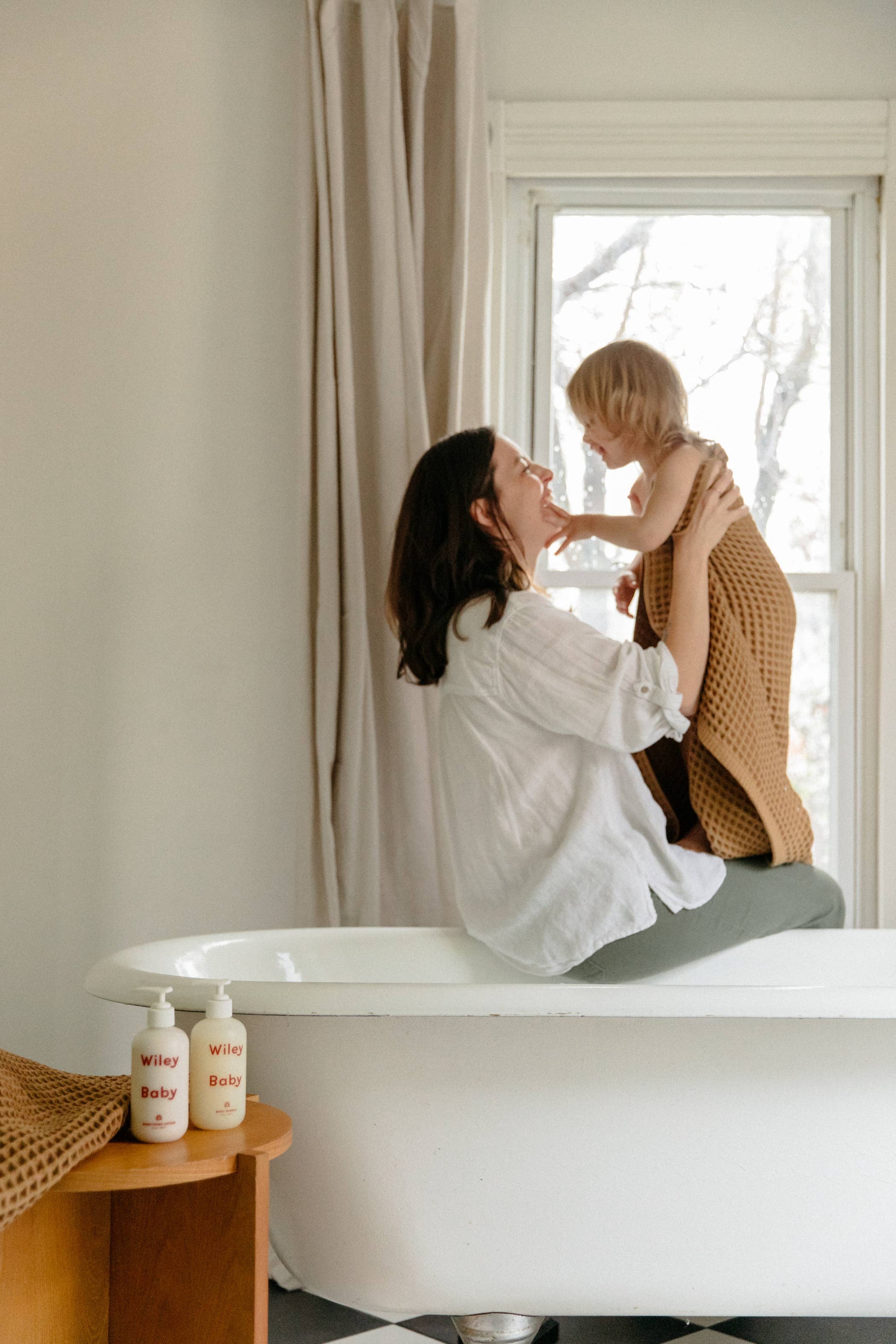 A woman wearing a white line shirt and grey trousers holds a child who has a brown blanket wrapped around them, sitting on the edge of a white bath in front of a window. Next to the bath, on top of a brown table are two bottles of Wiley Baby products which both have white white pumps and say Wiley Baby in red. 