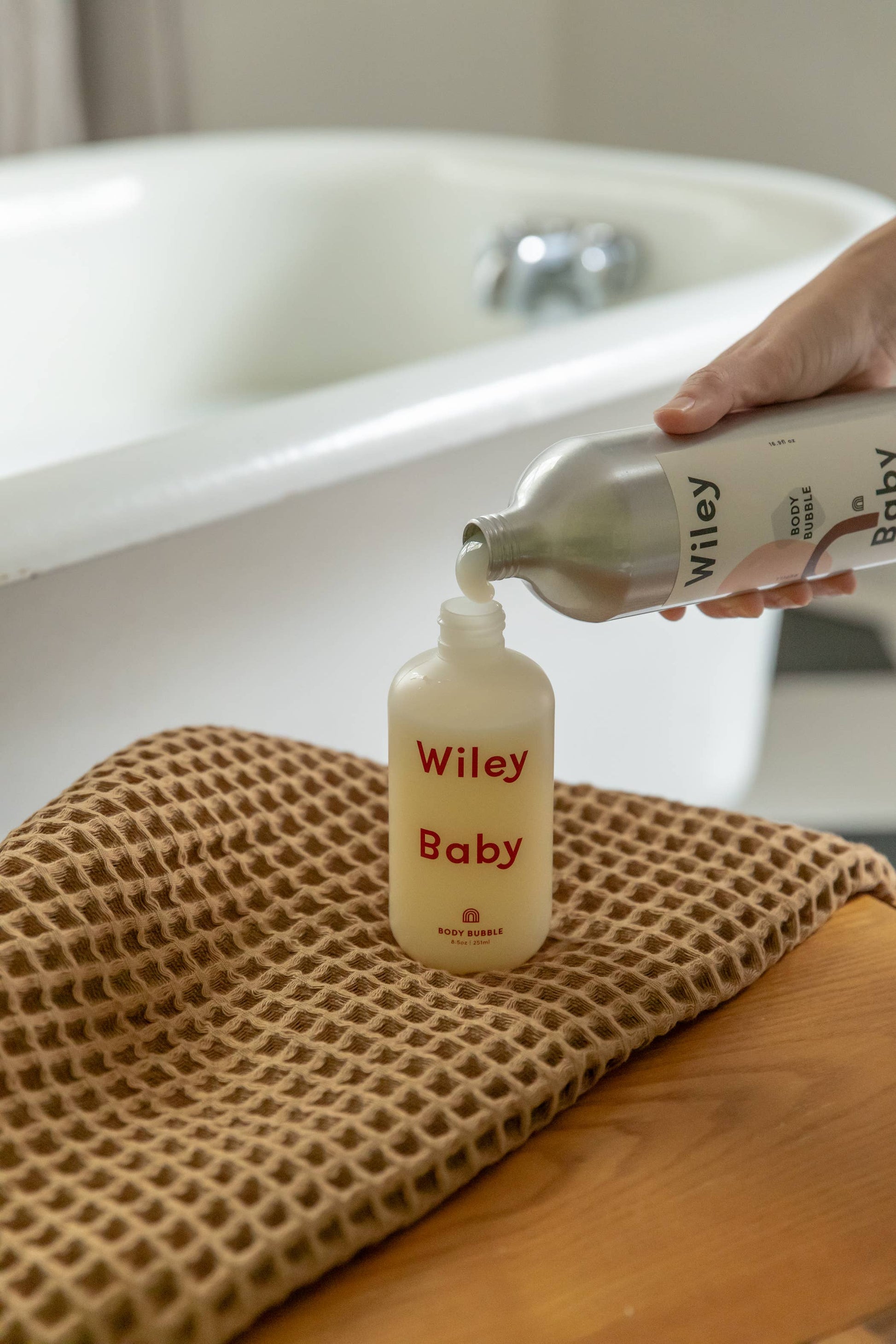 A metallic bottle of Wiley Baby body wash is being poured into a smaller, clear bottle of Wiley Baby body wash without its lid on. The bottle being refilled says Wiley Baby in red and is sitting on top of a brown cloth, on top of a brown table with a bathtub in the background. The metallic bottle has a white sticker on it that say Wiley at the top in black and Baby at the bottom with pink and brown abstract shapes around the letters. 