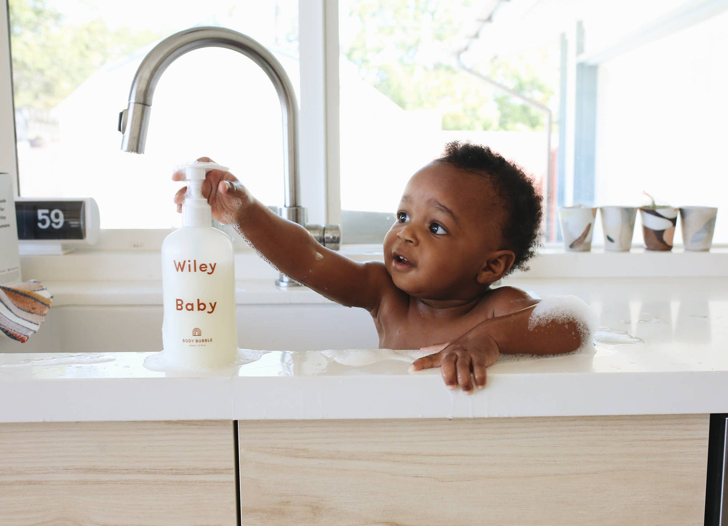 A baby sits inside of a large white kitchen sink with a large grey tap in front of a window with a digital clock and cups in front of the window on a shelf. The baby has bubbles on their arm and is holding onto the pump of a bottle of Wiley Baby Body Bubble that says Wiley Baby in a red/brown colour. 