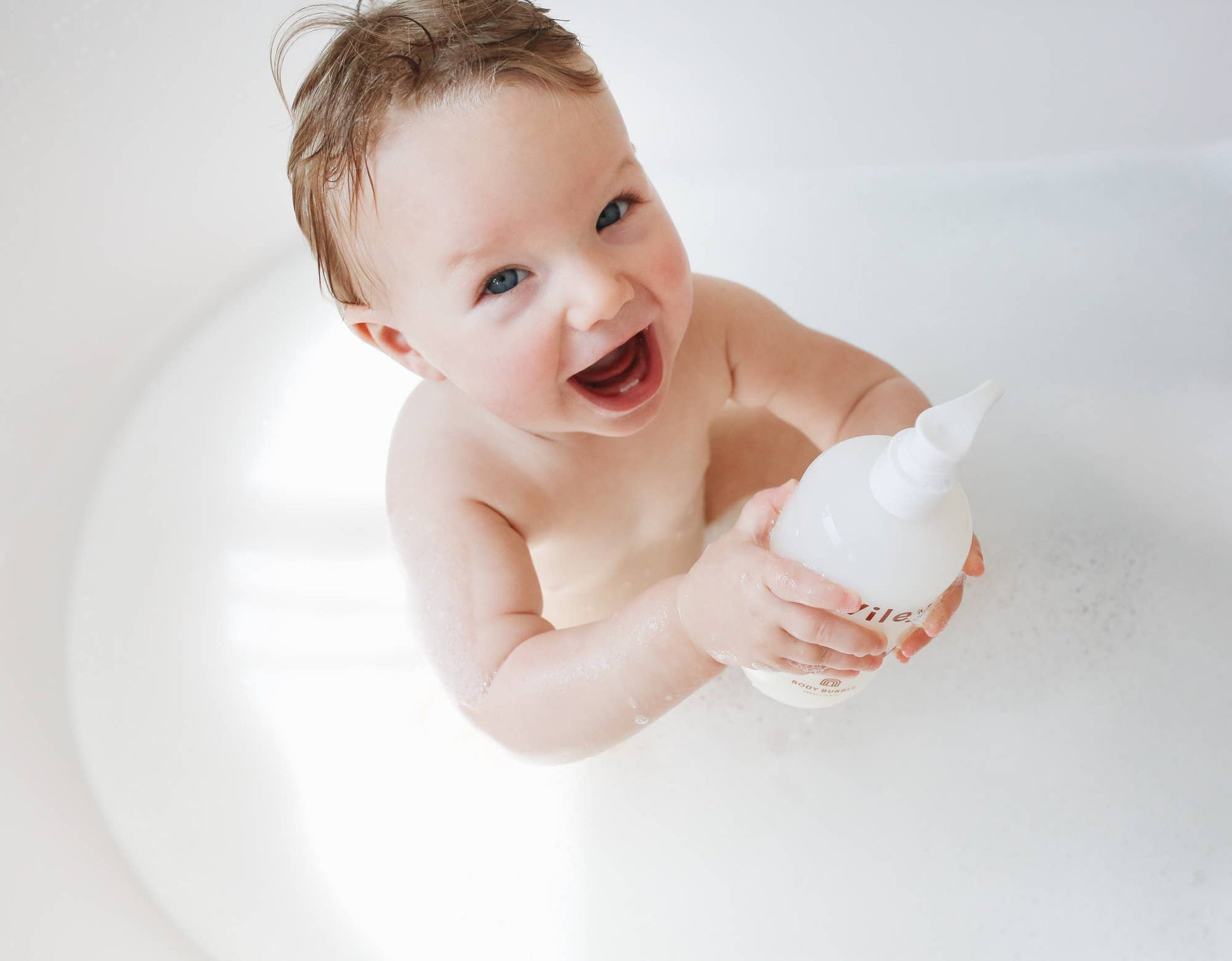 A overhead picture of a baby inside a bath holding a bottle of a Wiley Baby product that has a pump lid. 