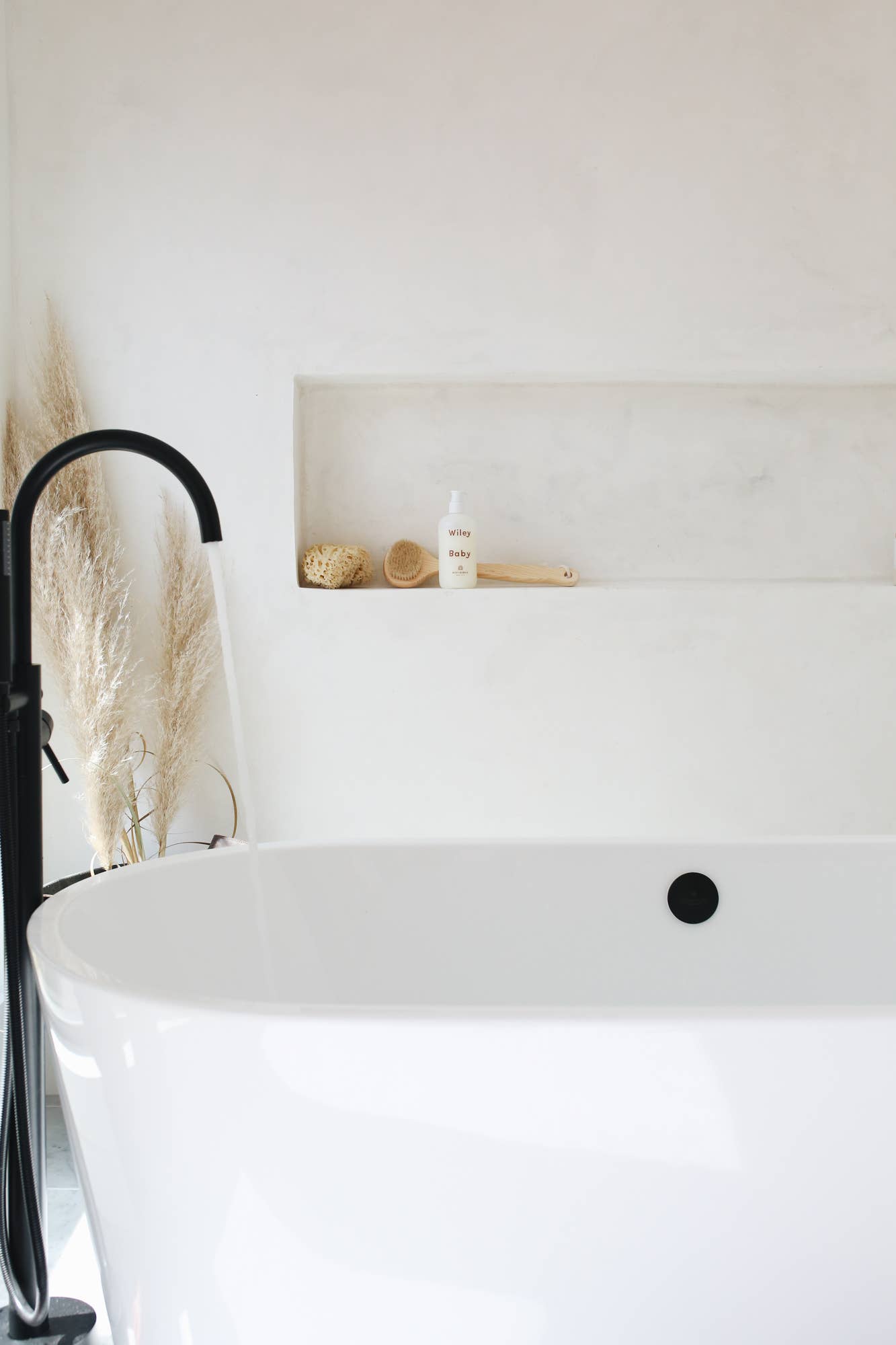 A bottle of the Wiley Baby Body Bubble sits inside a ledge next to a cream loofah and what appears to be a loofah on a wooden stick above a large white circular bathtub with a black tap and cream feather flowers next to it. 