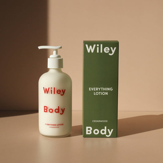 A clear bottle with a white 
pump with the Wiley Body Everything Lotion inside, making the bottle appear white. In red letters it says Wiley Body and beneath that it says Everything Lotion. Next to the bottle is the packaging which is a dark green box that at the top in white says Wiley and at the bottom says Body. In this middle it says Everything Lotion and underneath that it says Cedarwood. This is against a pink background.