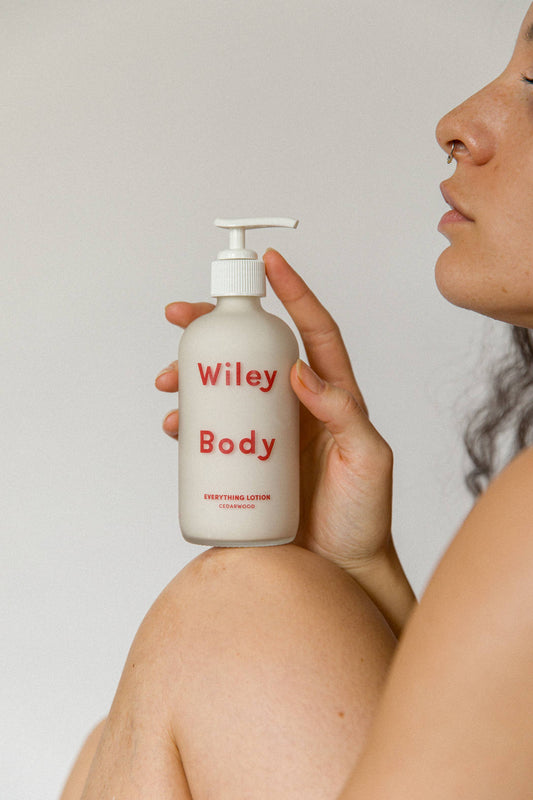 A bottle of the Wiley Body Everything Lotion , which is clear with a white liquid inside and says Wiley Body Everything Lotion in red is being held on top of someones knee with their side profile also in the image.