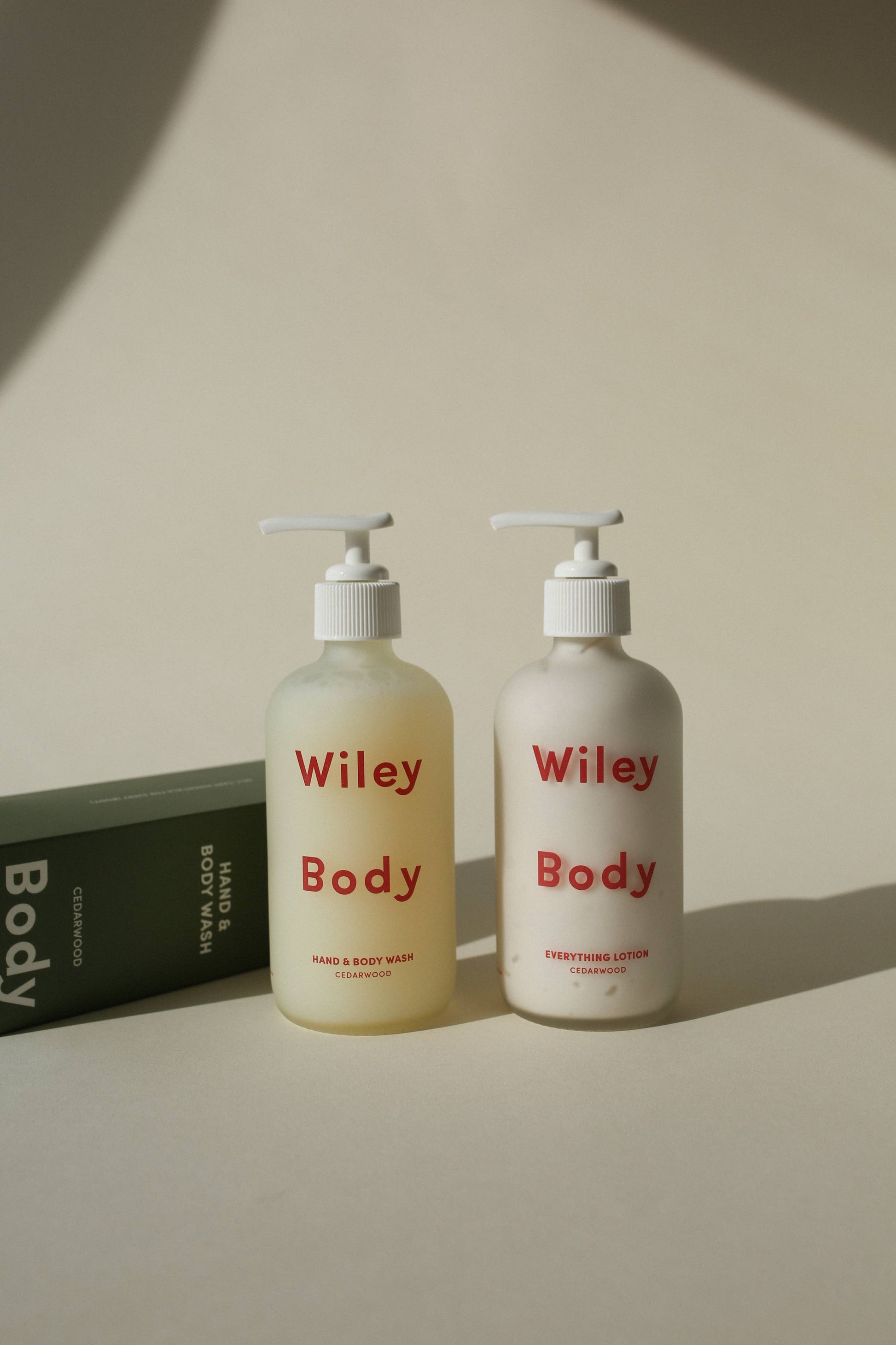 A bottle of the Wiley Body Hand And Body Wash stands next to a bottle of the Wiley Body Everything Lotion which is written ion both bottles in red. The Hand and Body Wash bottle appears yellow and the Everything Lotion appears white. This is against a white background and next to the Hand and Body Wash is its green packaging box which says Wiley at the top in white and Body at the bottom. 