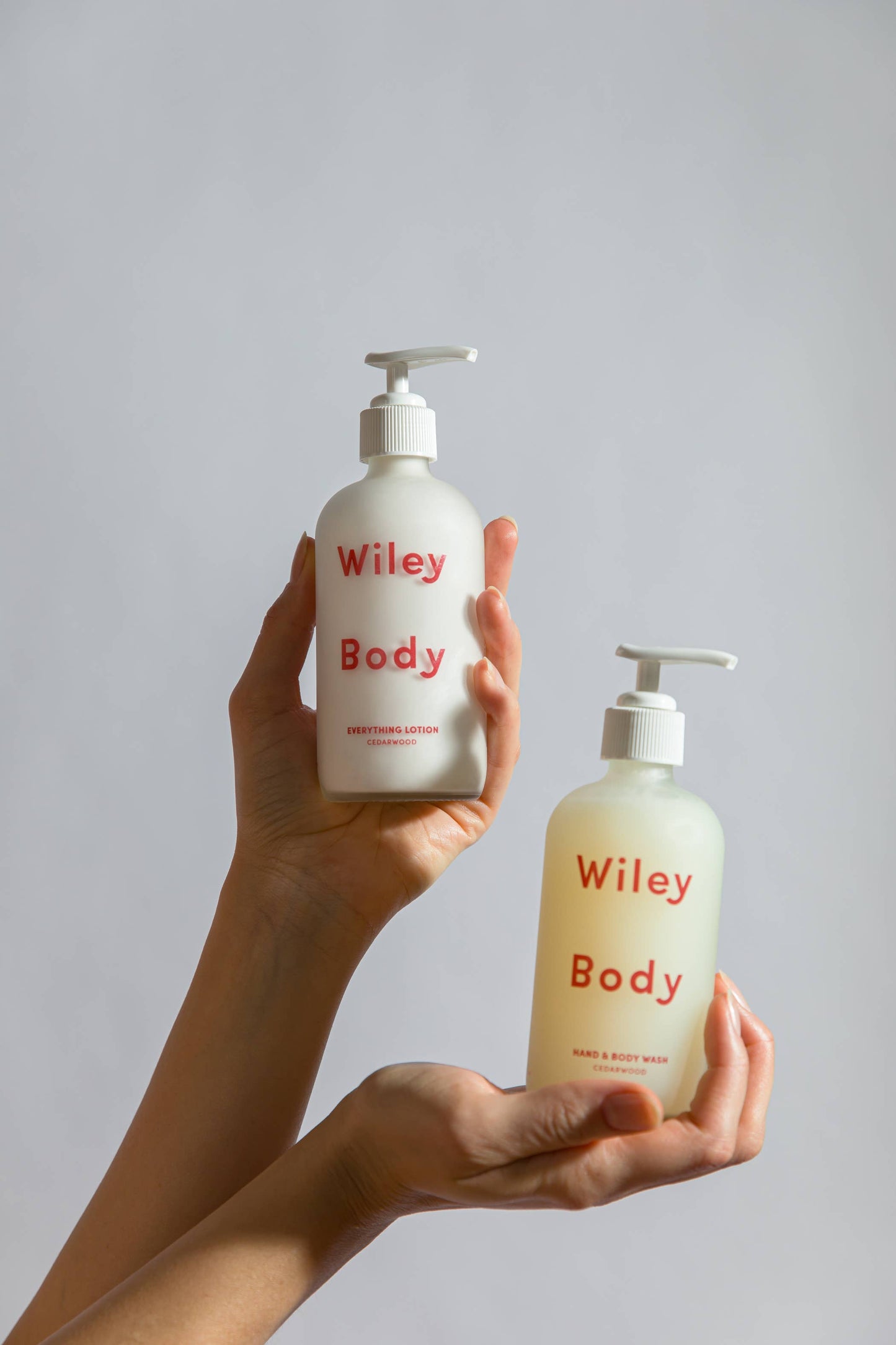 In either hands, up against a white background, someone is holding a bottle of the Wiley Body cream which has a white pump and appears to be white and says Wiley Body Everything Lotion in red. In the other hand they are holding the Wiley Body Hand and Body was which appears to be yellows and says Wiley Body Hand and Body Wash in red. 