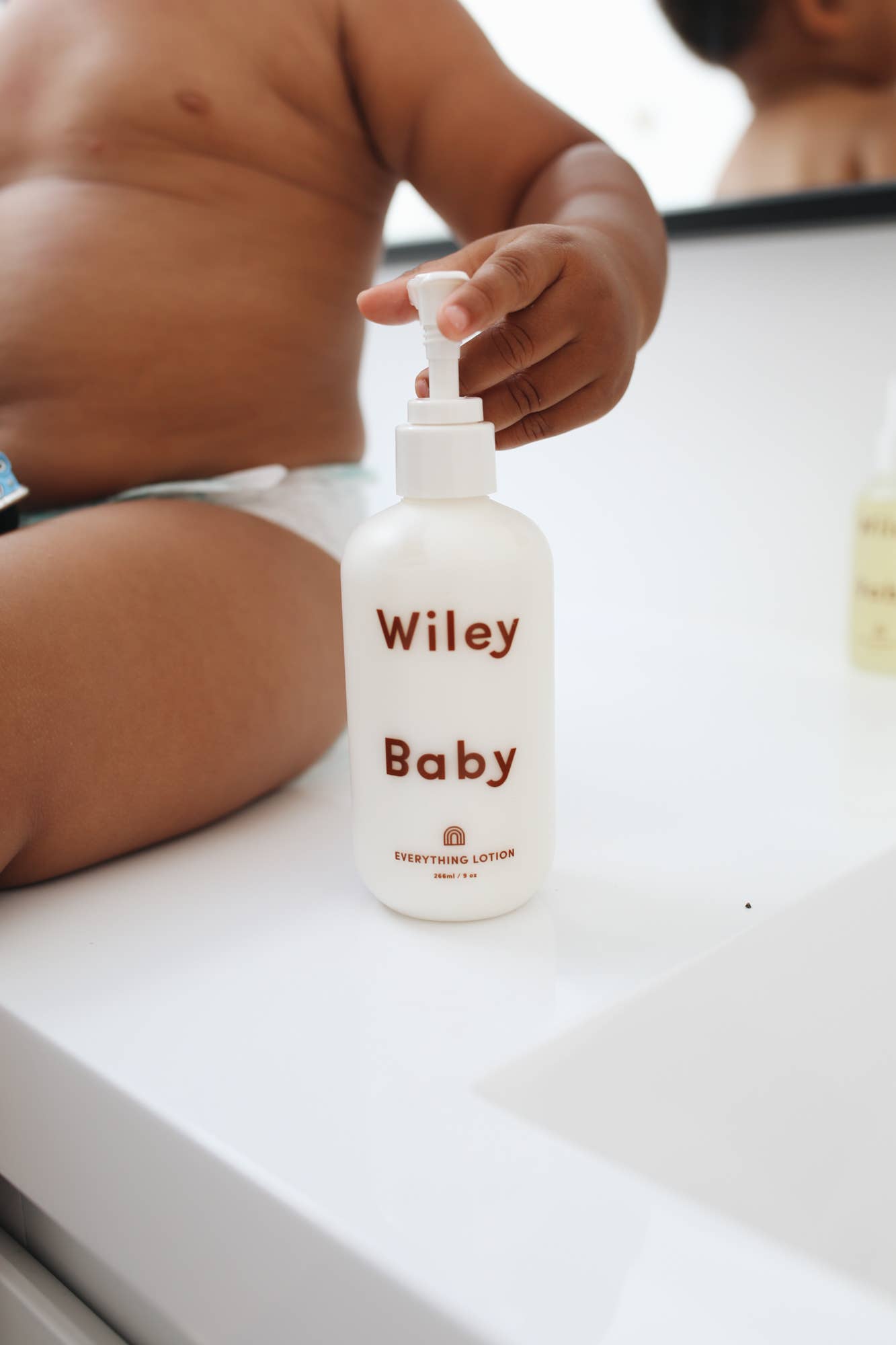 Wiley body everything lotion for baby, in a white cylindrical matte glass bottle with curved edges and a white pump cap, spelling Wiley baby in red text across the bottle, being touched by a baby in a nappy on a white marble countertop