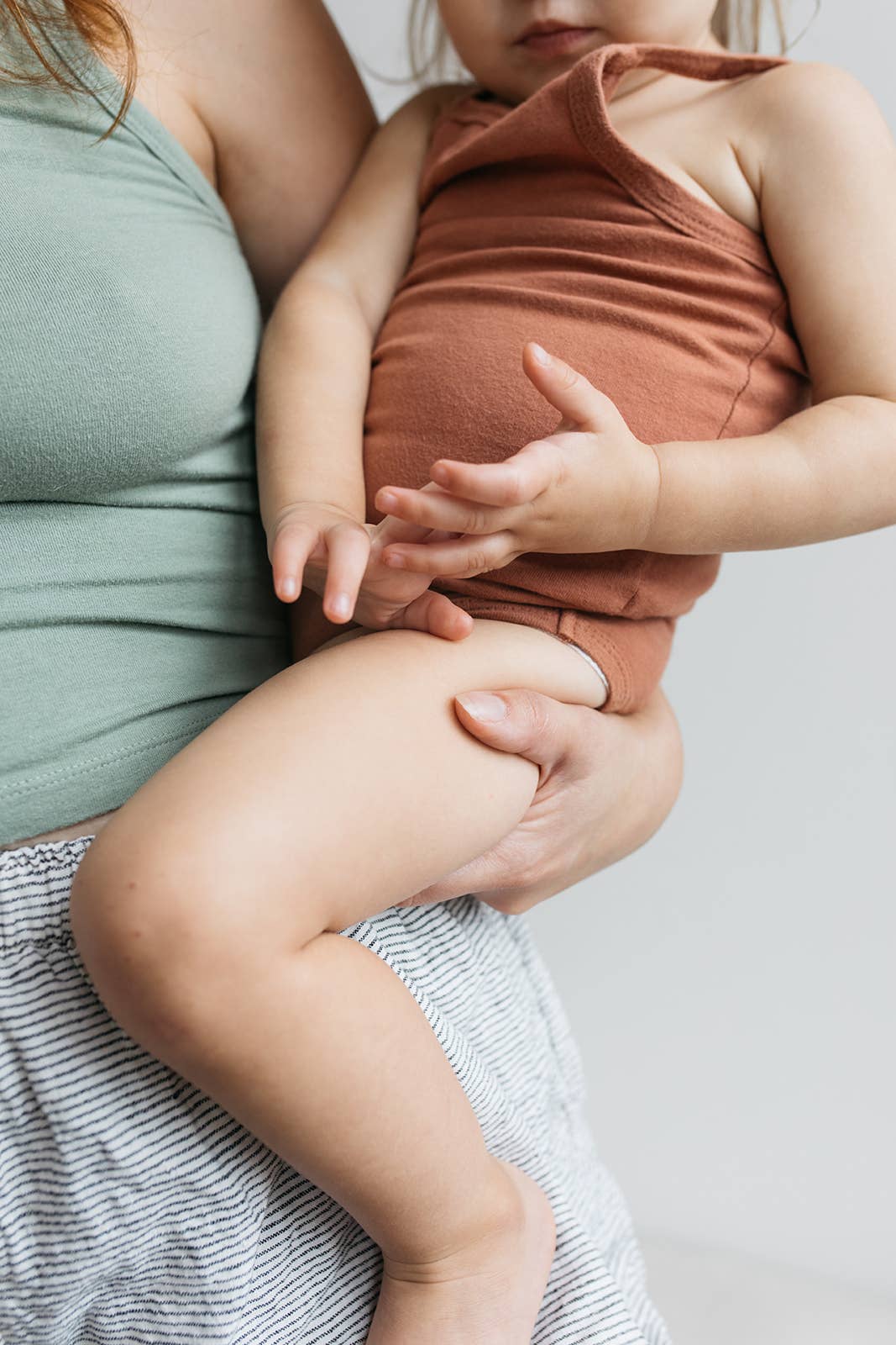 A close up of a woman in a green vest and striped pyjama pants holding a baby on her hips in her hand. The baby is wearing a rust coloured baby grow