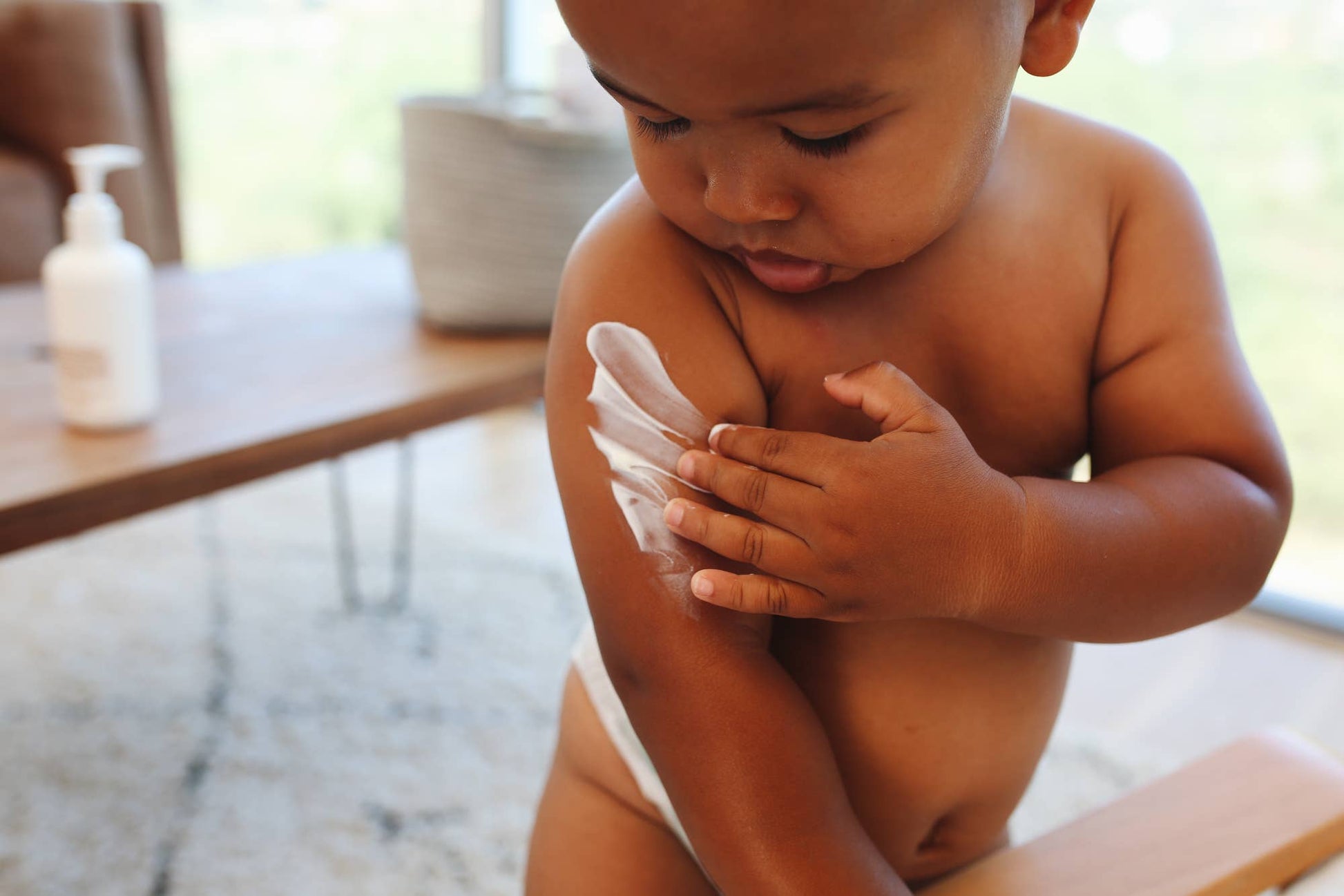 A baby infront of a wood table with the Wiley body everything lotion bottle on. The baby is spreading a patch of the white lotion across its arm, and is studying his actions