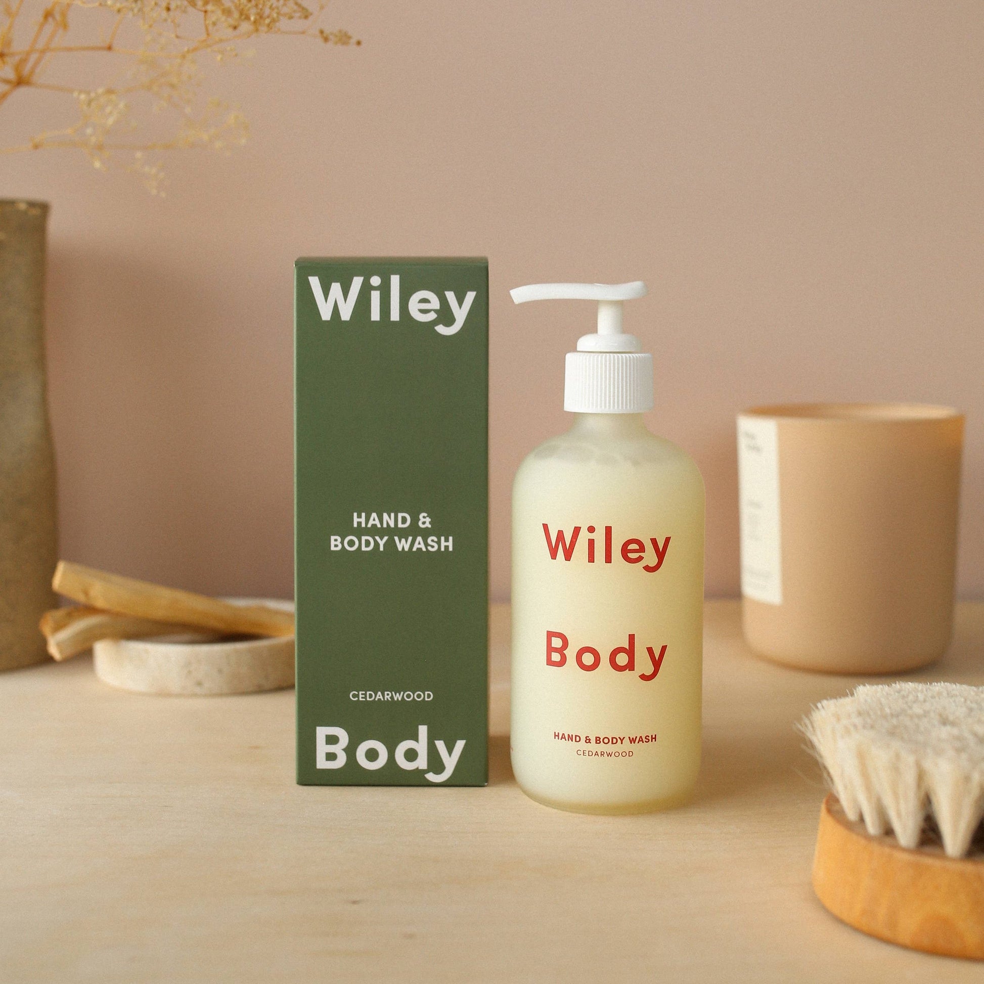 Wiley body hand and body wash in a clear matte cylindrical glass bottle with a white pump cap, on the front it has Wiley body written in large red lettering. Next to its packaging a dark green cuboid cardboard box with white text on it saying Wiley body at the top and bottom then smaller text saying hand and body wash. The background is mauve with a dry brush, a candle, and stone coaster with a sponge in the background