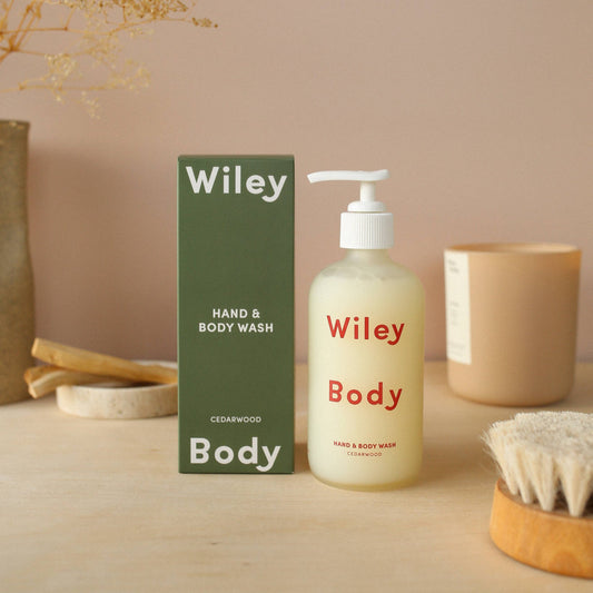 Wiley body hand and body wash in a clear matte cylindrical glass bottle with a white pump cap, on the front it has Wiley body written in large red lettering. Next to its packaging a dark green cuboid cardboard box with white text on it saying Wiley body at the top and bottom then smaller text saying hand and body wash. The background is mauve with a dry brush, a candle, and stone coaster with a sponge in the background