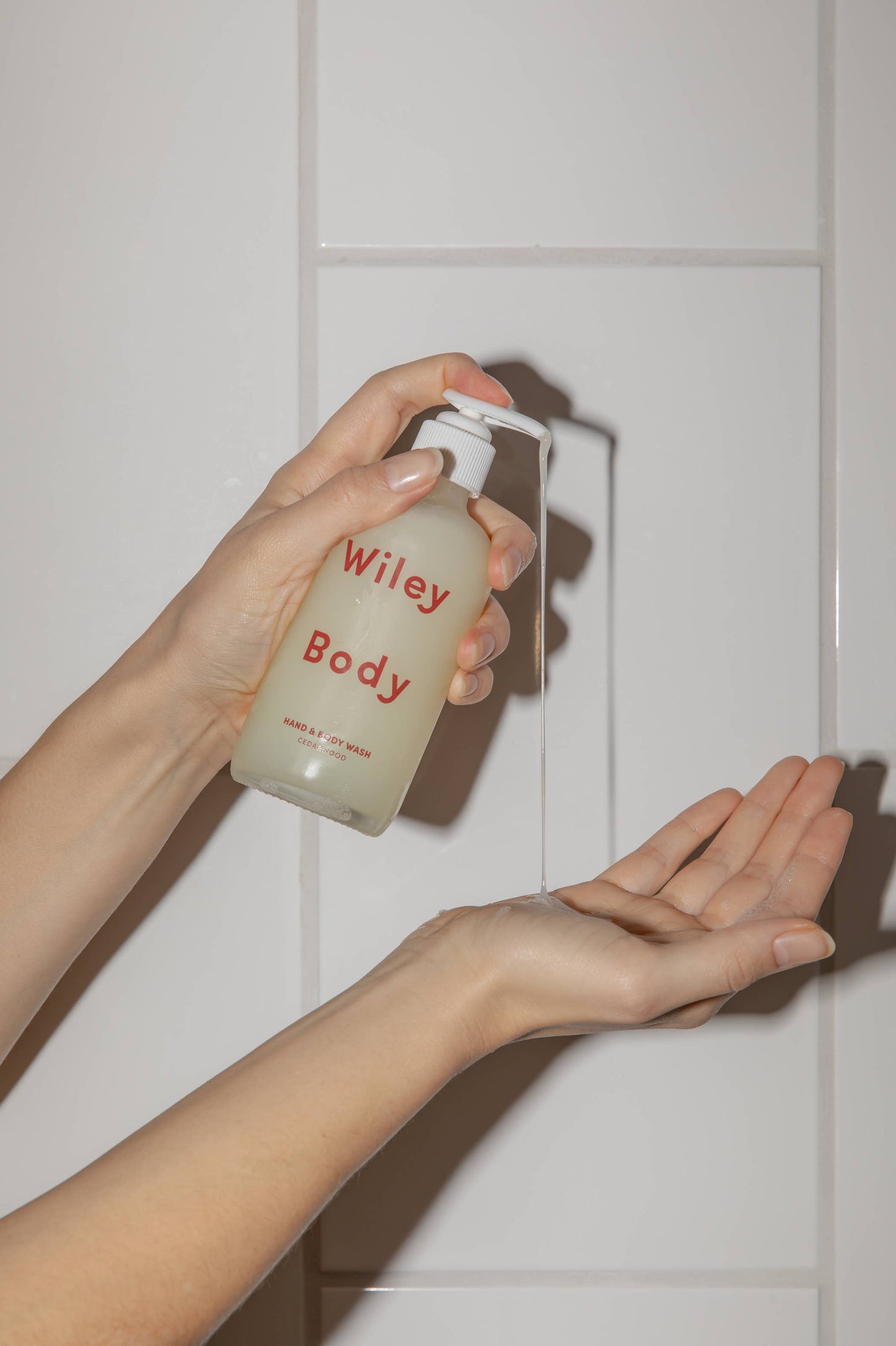 A woman’s hands in action pumping the Wiley body hand and body wash into her palm, in front of white tile