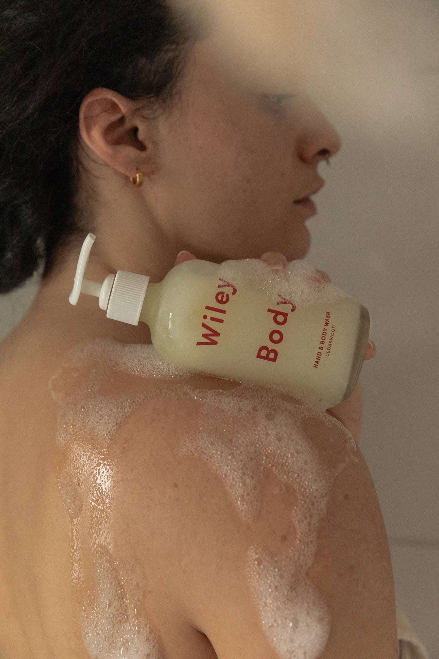 A woman holding the Wiley body hand and body wash bottle in her hand with suds around the bottle and her shoulder.