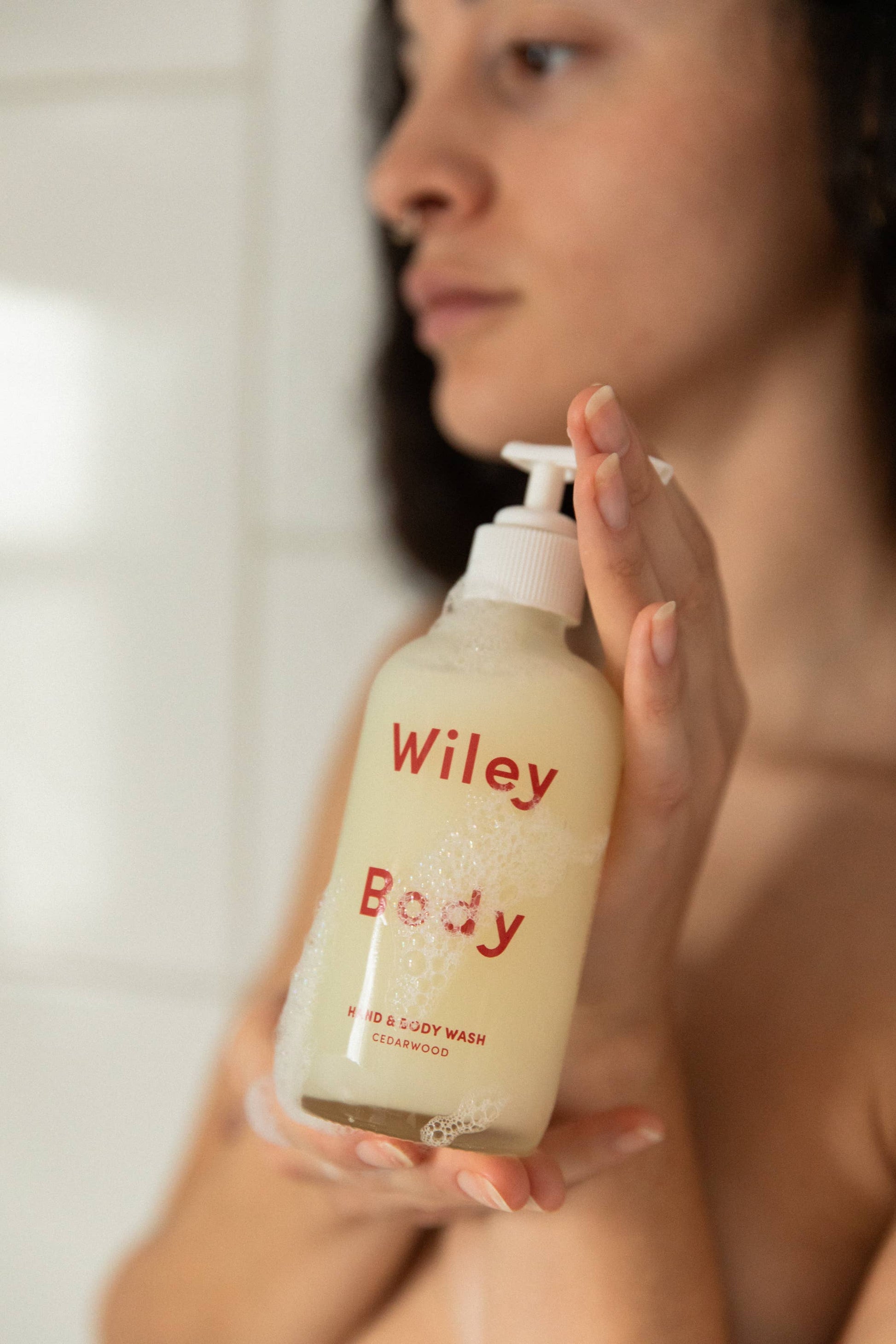 A woman caressing the Wiley body hand and body wash bottle in her hands, in front of a tiled wall. 