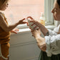 A brown haired woman holding the Wiley body everything lotion for baby, in a white cylindrical matte glass bottle with curved edges and a white pump cap, spelling Wiley baby in red text across the bottle, she is pumping some lotion on to a child’s hands in front of a window. The woman is wearing a rolled up white shirt and the child has their hand out stretched wearing a rush coloured vest and shorts