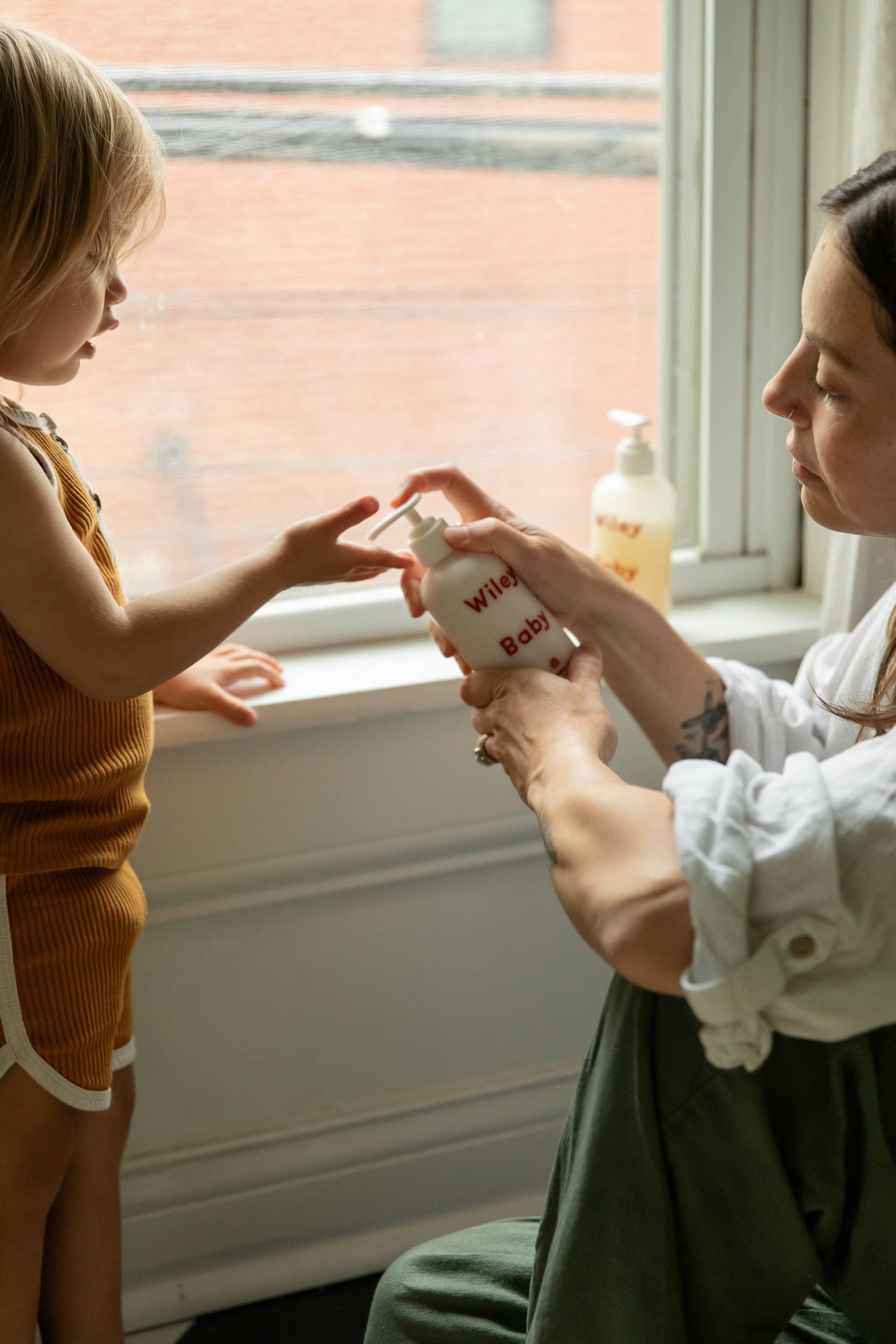 A brown haired woman holding the Wiley body everything lotion for baby, in a white cylindrical matte glass bottle with curved edges and a white pump cap, spelling Wiley baby in red text across the bottle, she is pumping some lotion on to a child’s hands in front of a window. The woman is wearing a rolled up white shirt and the child has their hand out stretched wearing a rush coloured vest and shorts