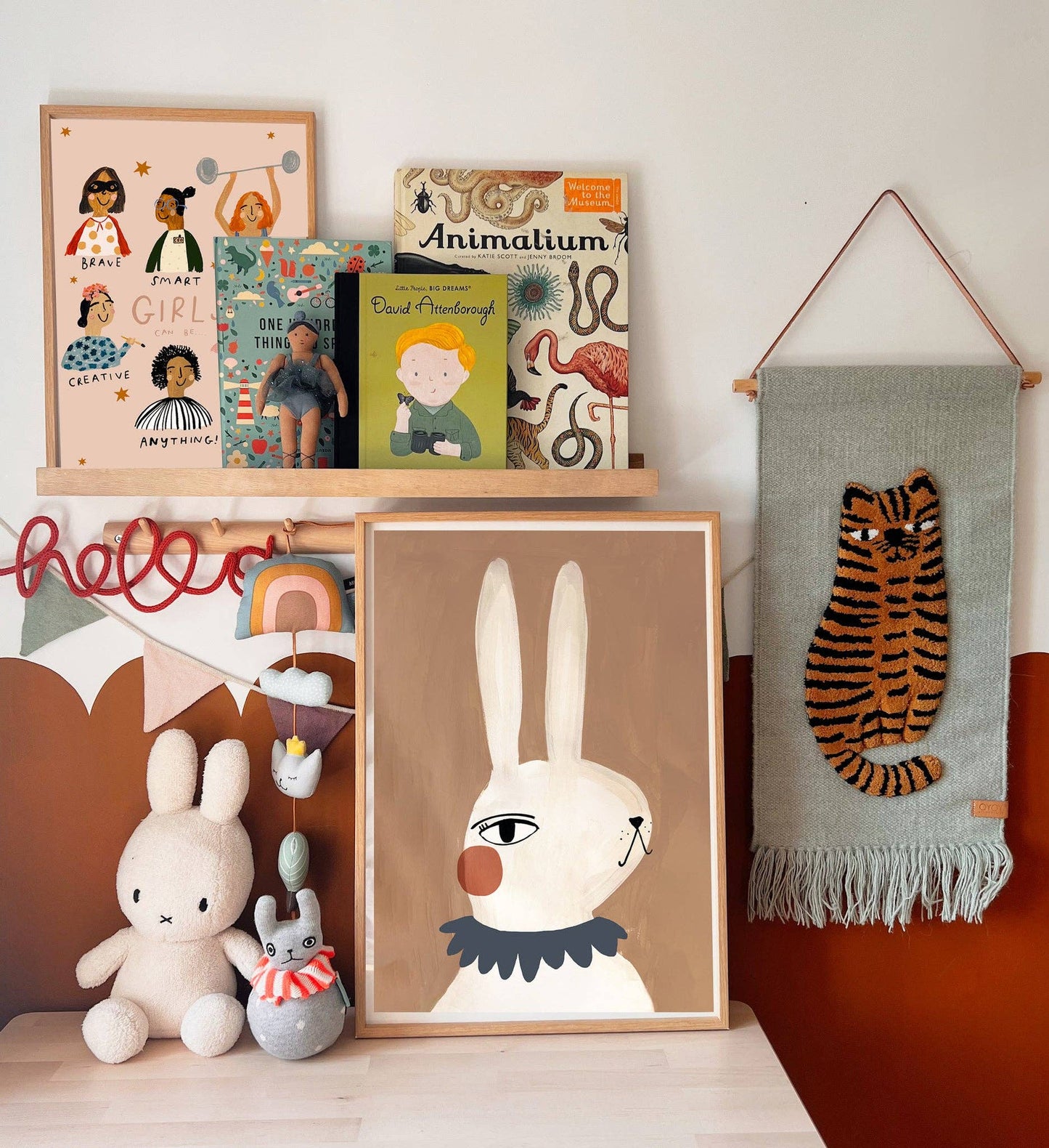 Hand-painted Mrs Rabbit print with rosy cheeks, designed for nursery decor, printed on high-quality 250gsm matte paper, featuring a friendly rabbit illustration. The print sits in a wooden frame, standing alongside some cuddly rabbit toys, some books, and a a tiger wall hanging