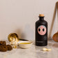 A matte black bottle of Yiayia and Friends Extra Virgin Olive Oil with a playful illustrated face, surrounded by garlic, golden measuring spoons, dark bread, and a small dish of olive oil on a marble surface.