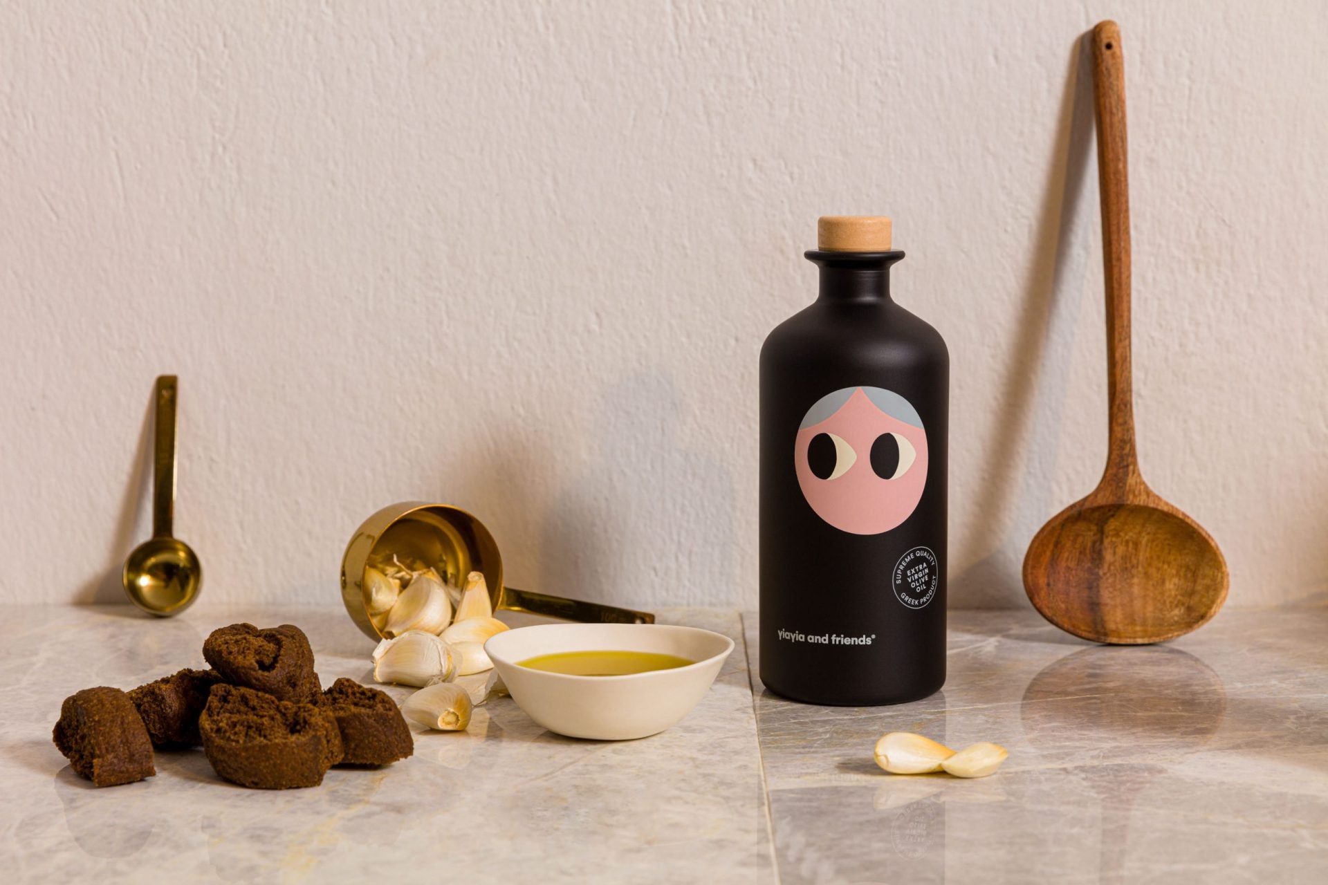 A matte black bottle of Yiayia and Friends Extra Virgin Olive Oil with a playful illustrated face, surrounded by garlic, golden measuring spoons, dark bread, and a small dish of olive oil on a marble surface.