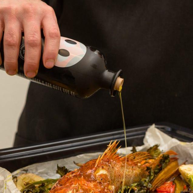  person pouring Yiayia and Friends Extra Virgin Olive Oil from a black bottle onto a whole roasted fish with vegetables, highlighting its premium quality and culinary use.