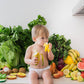 Baby sitting on a kitchen floor surrounded by fruits and vegetables