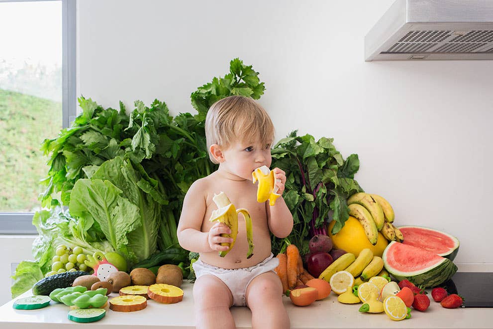 Baby sitting on a kitchen floor surrounded by fruits and vegetables