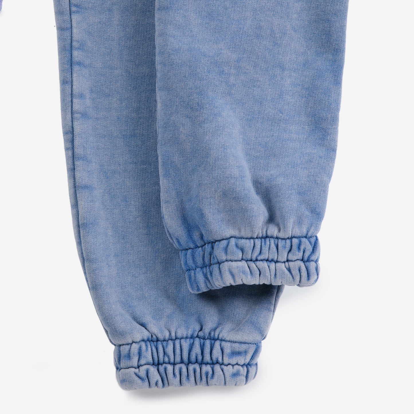 A close-up of the lower legs of a pair of washed blue sweatpants, highlighting the elasticated cuffs. The fabric has a soft, slightly faded look, and the elastic gathering creates a gently ruched effect. The stitching details along the seams are visible, enhancing the relaxed, vintage-inspired aesthetic.