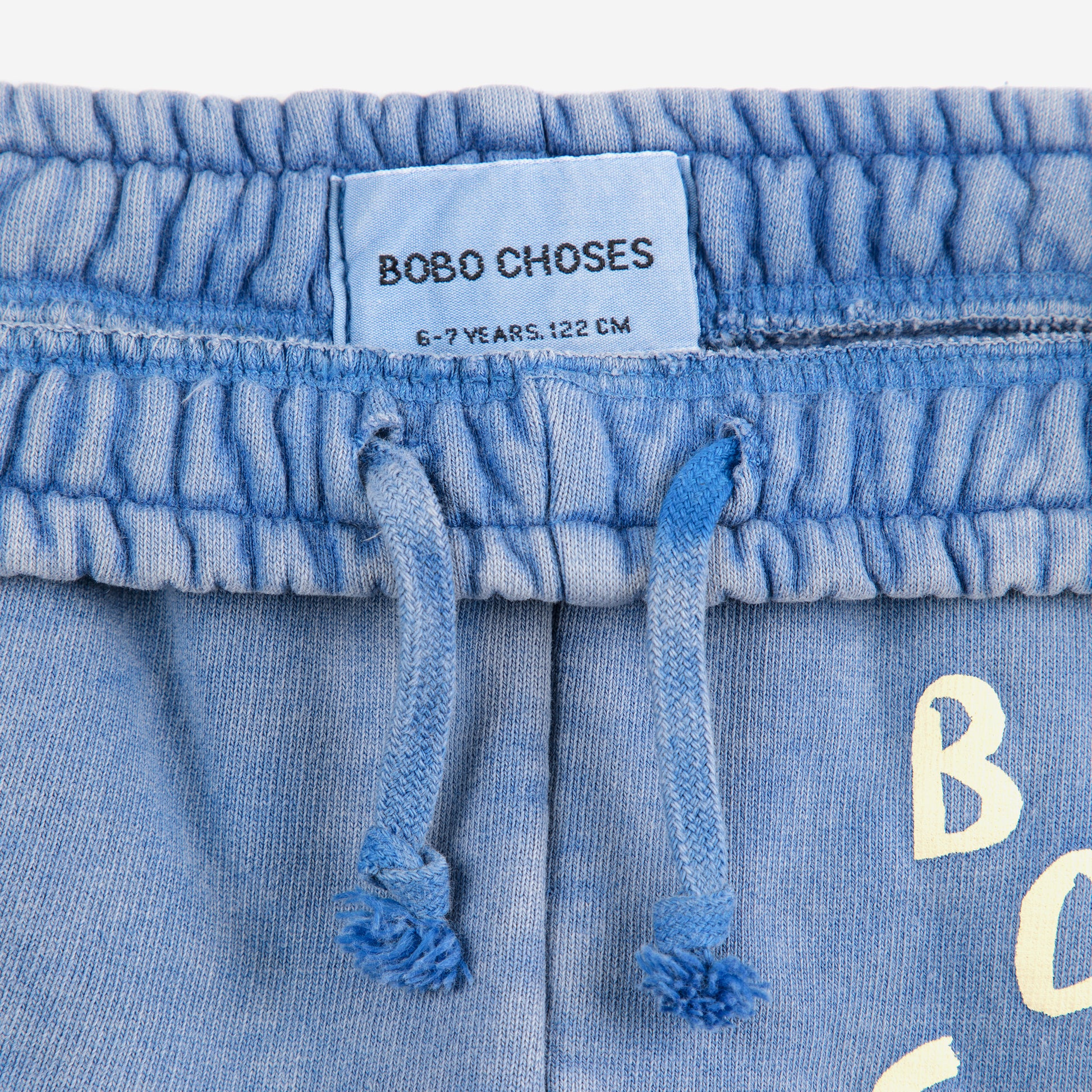 A close-up of the waistband of a pair of washed blue sweatpants, featuring an elasticated band with a gathered texture and a matching drawstring with knotted and frayed ends. A light blue fabric label sewn inside the waistband displays the brand name "BOBO CHOSES" along with the size "6-7 YEARS, 122 CM." Part of the "BOBO CHOSES" logo print is visible on the right side.