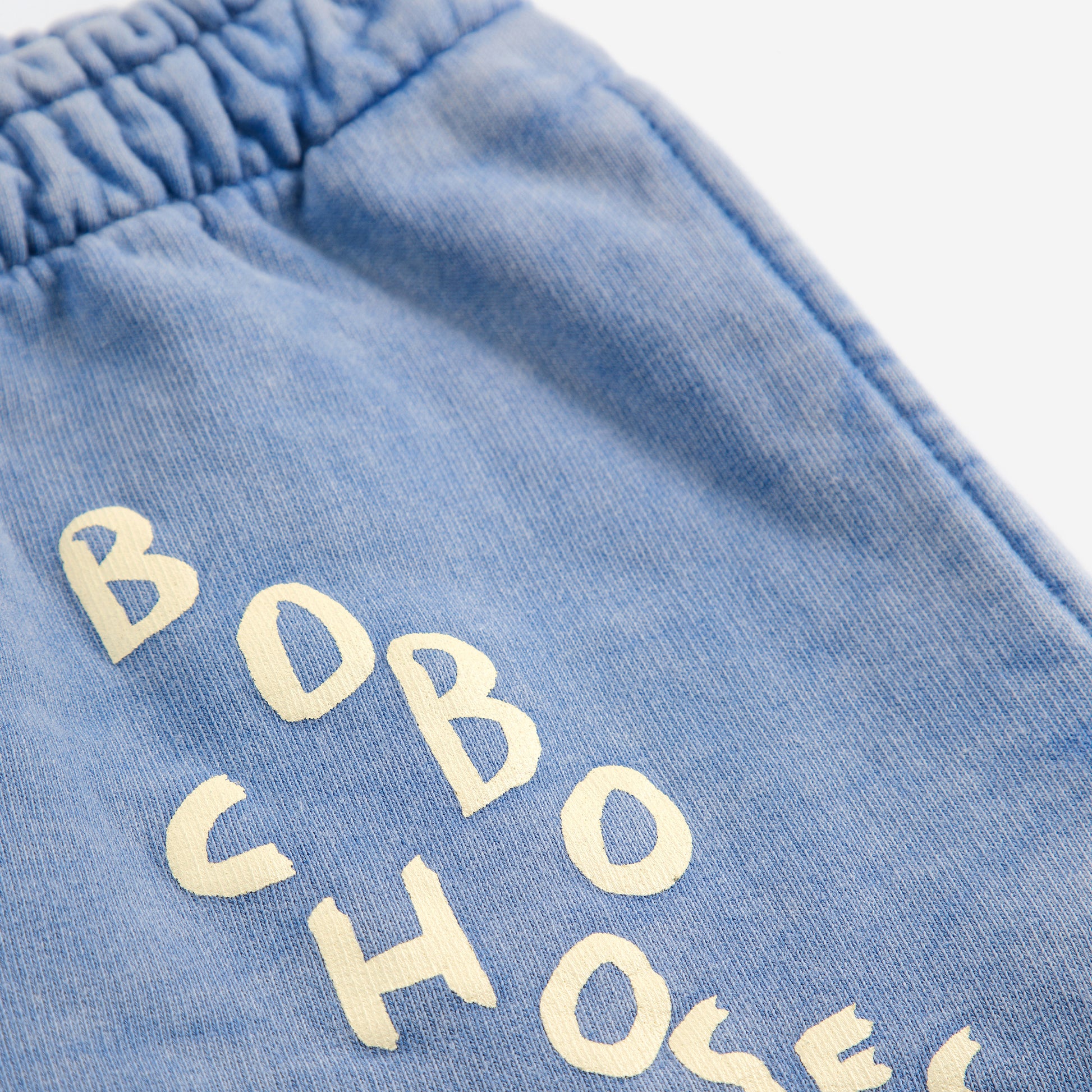 A close-up of the upper left leg of a pair of washed blue sweatpants, showcasing the "BOBO CHOSES" logo printed in an irregular, hand-drawn style in off-white. The elasticated waistband is partially visible, along with subtle stitching details. The fabric has a soft, slightly faded texture.