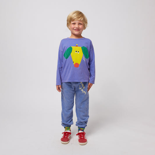 A young child with short blonde hair and a cheerful smile stands facing forward, wearing washed blue sweatpants with an elasticated waistband and cuffs. The sweatpants feature a "BOBO CHOSES" logo printed in an irregular, off-white font on the upper left leg. They are paired with a long-sleeved purple T-shirt featuring a large, abstract yellow dog graphic with green ears and a red nose. The child is also wearing colourful patterned socks and red sneakers with white soles. The background is plain white.