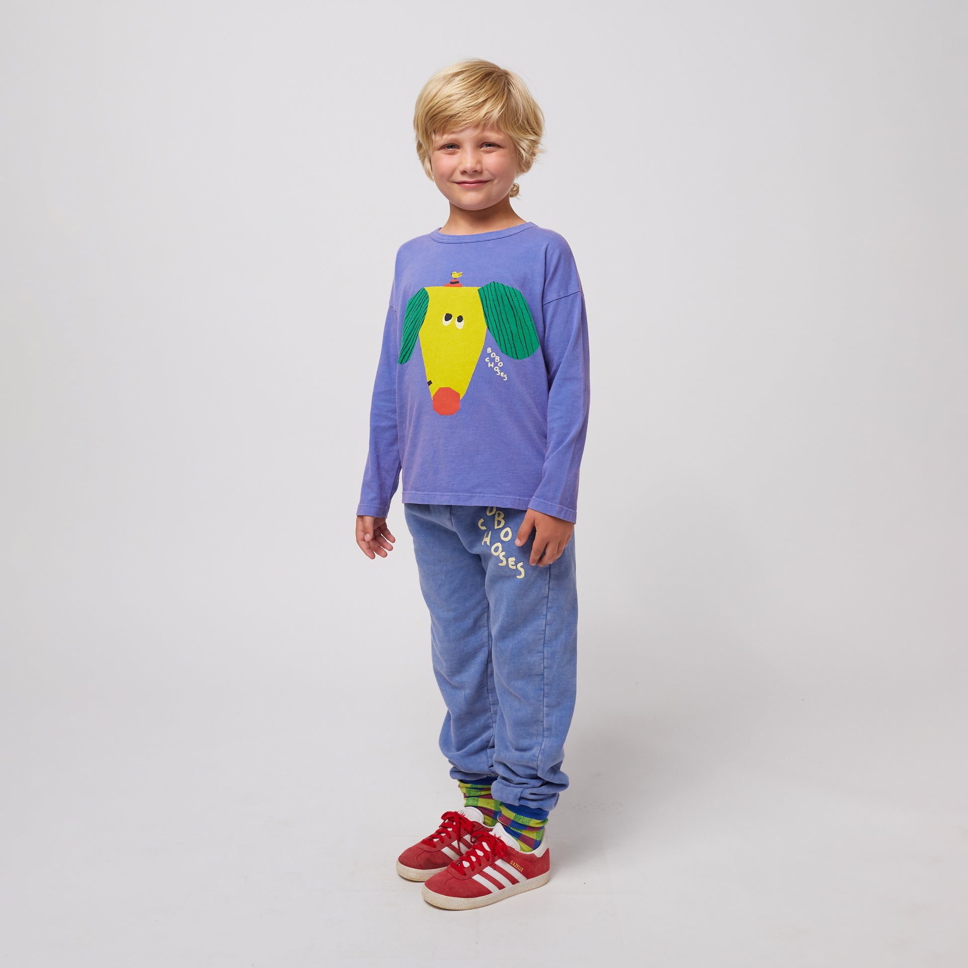 A young child with short blonde hair and a happy expression stands slightly turned to the side, wearing washed blue sweatpants with an elasticated waistband and cuffs. The sweatpants feature a "BOBO CHOSES" logo in an off-white, irregular font on the upper left leg. They are paired with a long-sleeved purple T-shirt featuring a large, abstract yellow dog graphic with green ears and a red nose. The child is also wearing colourful patterned socks and red Adidas sneakers with white stripes and soles. The backg