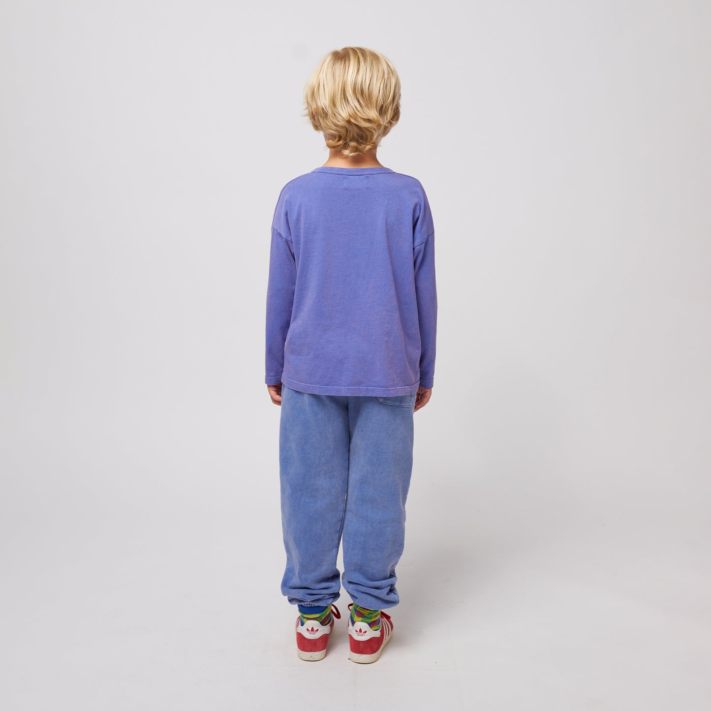 A young child with short blonde hair is seen from the back, wearing washed blue sweatpants with a relaxed fit, an elasticated waistband and cuffs, and a single square patch pocket on the right side. They are also wearing a long-sleeved purple T-shirt, colourful patterned socks, and red Adidas sneakers with white soles and the Adidas trefoil logo on the heel. The background is plain white.