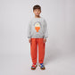 A young child stands facing forward, wearing bright orange sweatpants with visible stitched seams and a grey sweatshirt featuring a playful egg-shaped graphic with "BOBO CHOSES" text. They have a relaxed stance, their hands at their sides, and are wearing colourful sneakers with red, blue, and yellow straps over a chunky sole. The background is plain white.