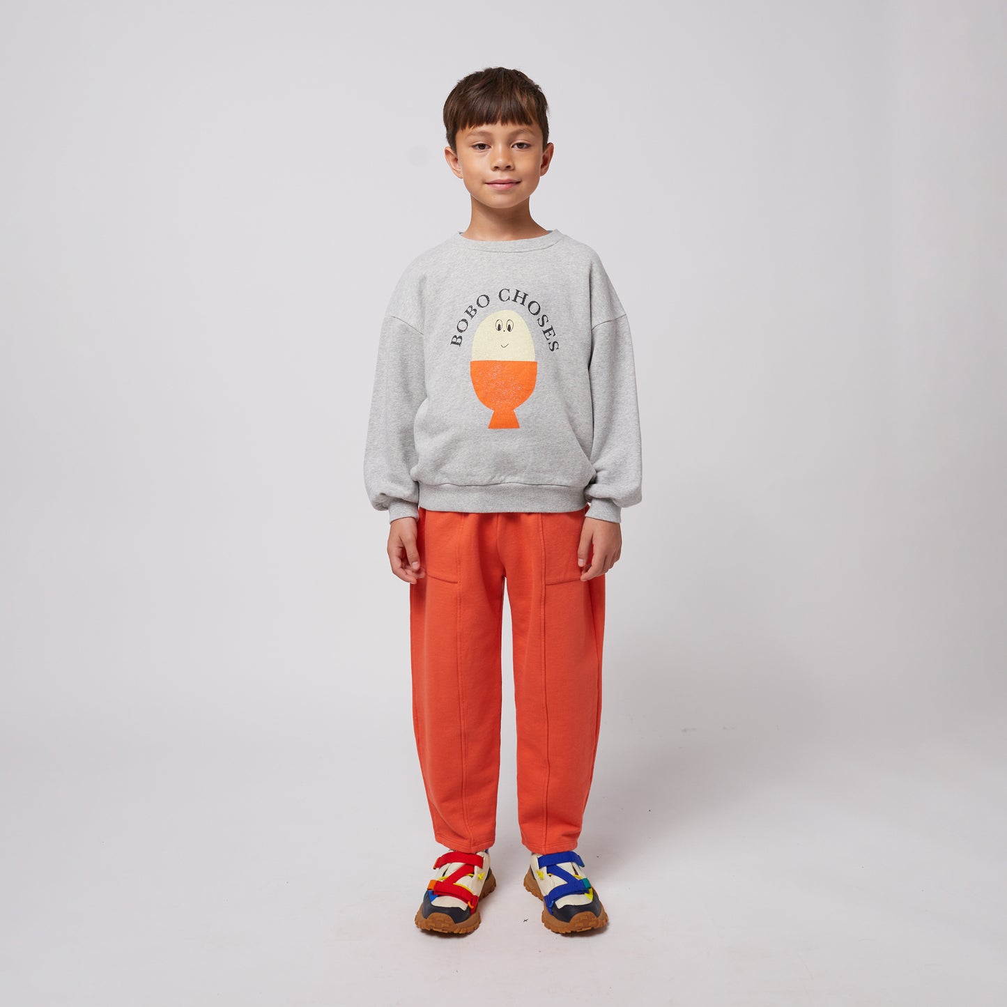 A young child stands facing forward, wearing bright orange sweatpants with visible stitched seams and a grey sweatshirt featuring a playful egg-shaped graphic with "BOBO CHOSES" text. They have a relaxed stance, their hands at their sides, and are wearing colourful sneakers with red, blue, and yellow straps over a chunky sole. The background is plain white.