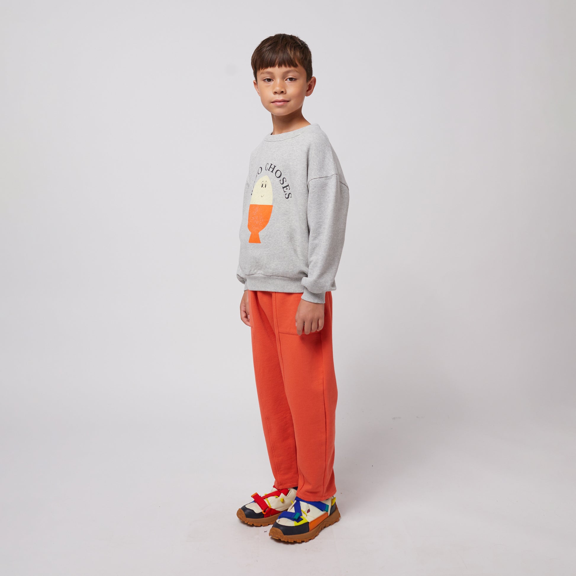 A young child stands slightly turned to the side, wearing bright orange sweatpants with stitched seam details and a grey sweatshirt featuring a playful egg-shaped graphic with "BOBO CHOSES" text. They have a neutral expression and wear colourful sneakers with red, blue, and yellow straps over a chunky sole. The background is plain white.