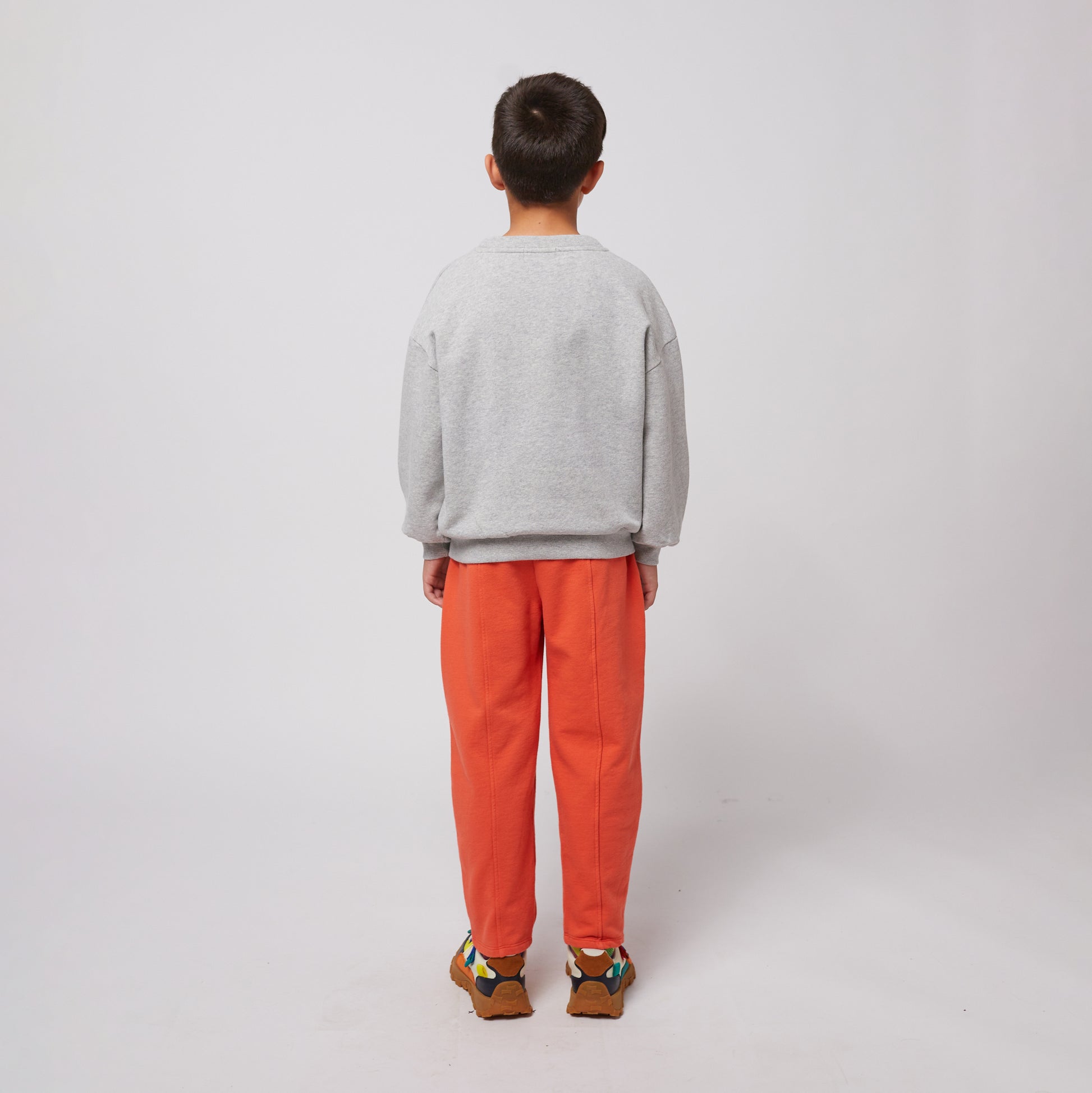 A young child is seen from the back, wearing bright orange sweatpants with stitched seams and a relaxed fit, paired with a plain grey sweatshirt. They have short, neatly trimmed hair and stand with a neutral posture. They are also wearing chunky sneakers with a multicoloured design featuring shades of blue, yellow, and brown. The background is plain white.