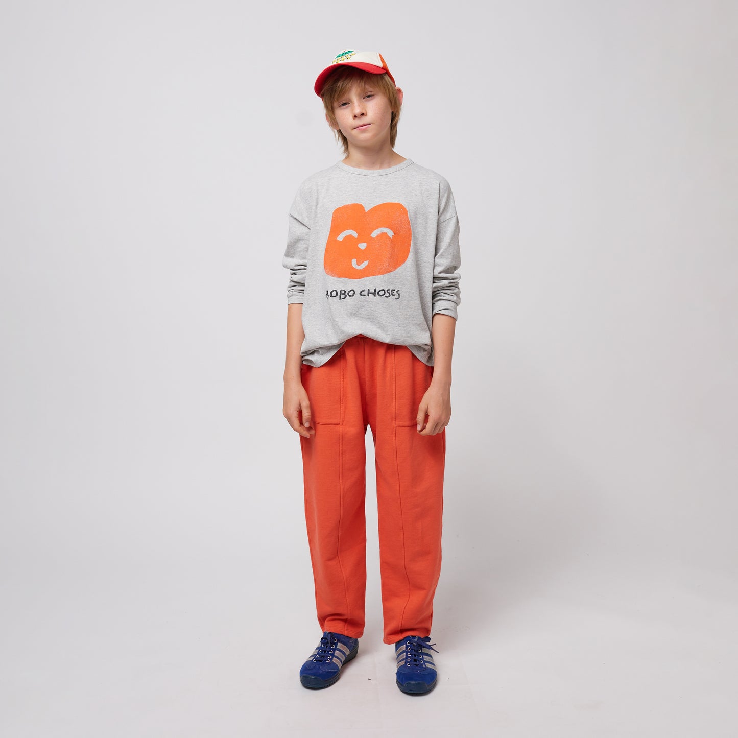 A young child stands facing forward, wearing bright orange sweatpants with stitched seam details and a grey sweatshirt featuring a playful orange smiling face graphic with "BOBO CHOSES" text. They are also wearing a red and white trucker cap and blue Adidas sneakers with white stripes. Their hands rest casually at their sides, and they have a relaxed, neutral expression. The background is plain white.