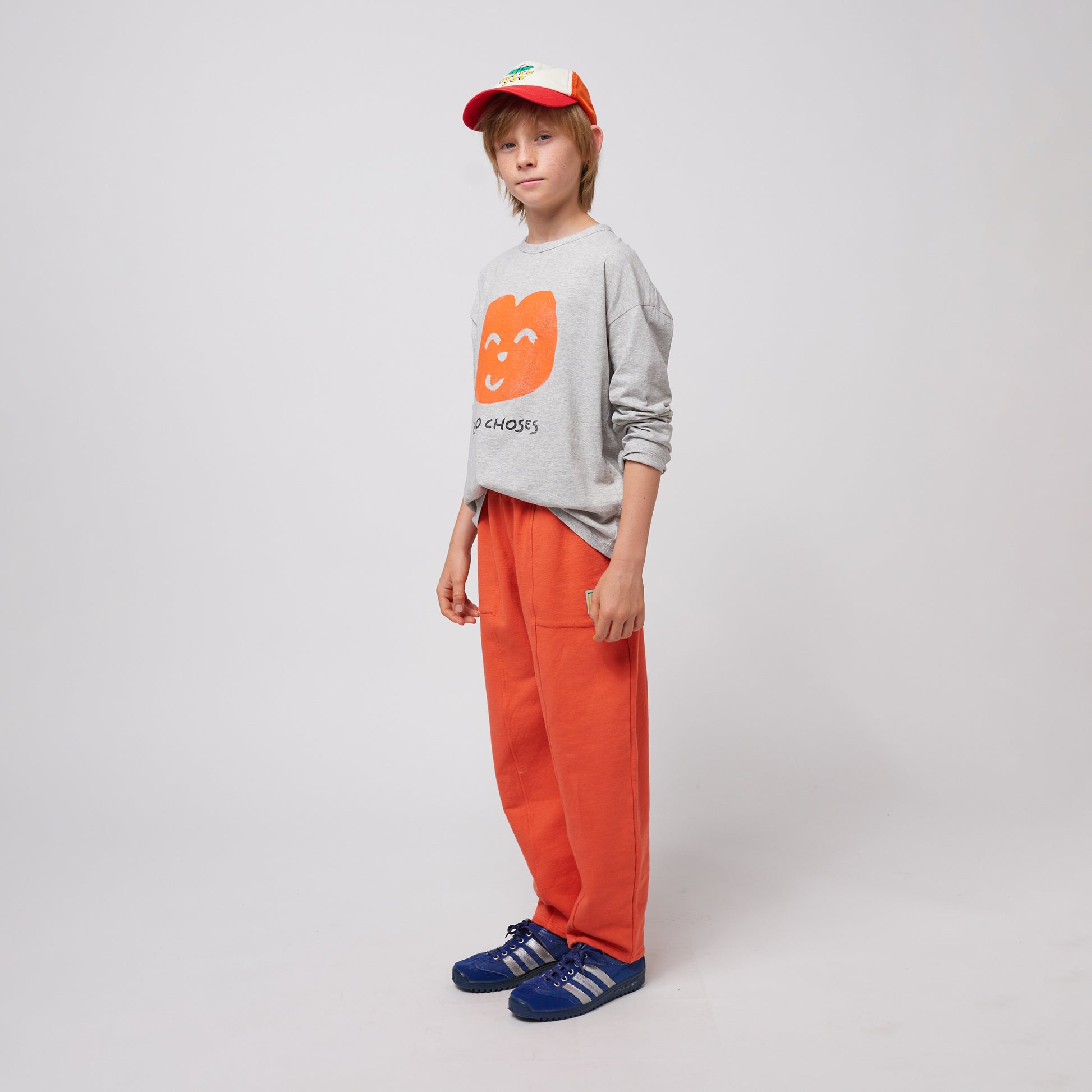 A young child stands slightly turned to the side, wearing bright orange sweatpants with visible stitched seams and a grey sweatshirt featuring a playful orange smiling face graphic with "BOBO CHOSES" text. They are also wearing a red and white trucker cap and blue Adidas sneakers with white stripes. Their expression is neutral, and their hand rests near a small side pocket with a logo patch. The background is plain white.