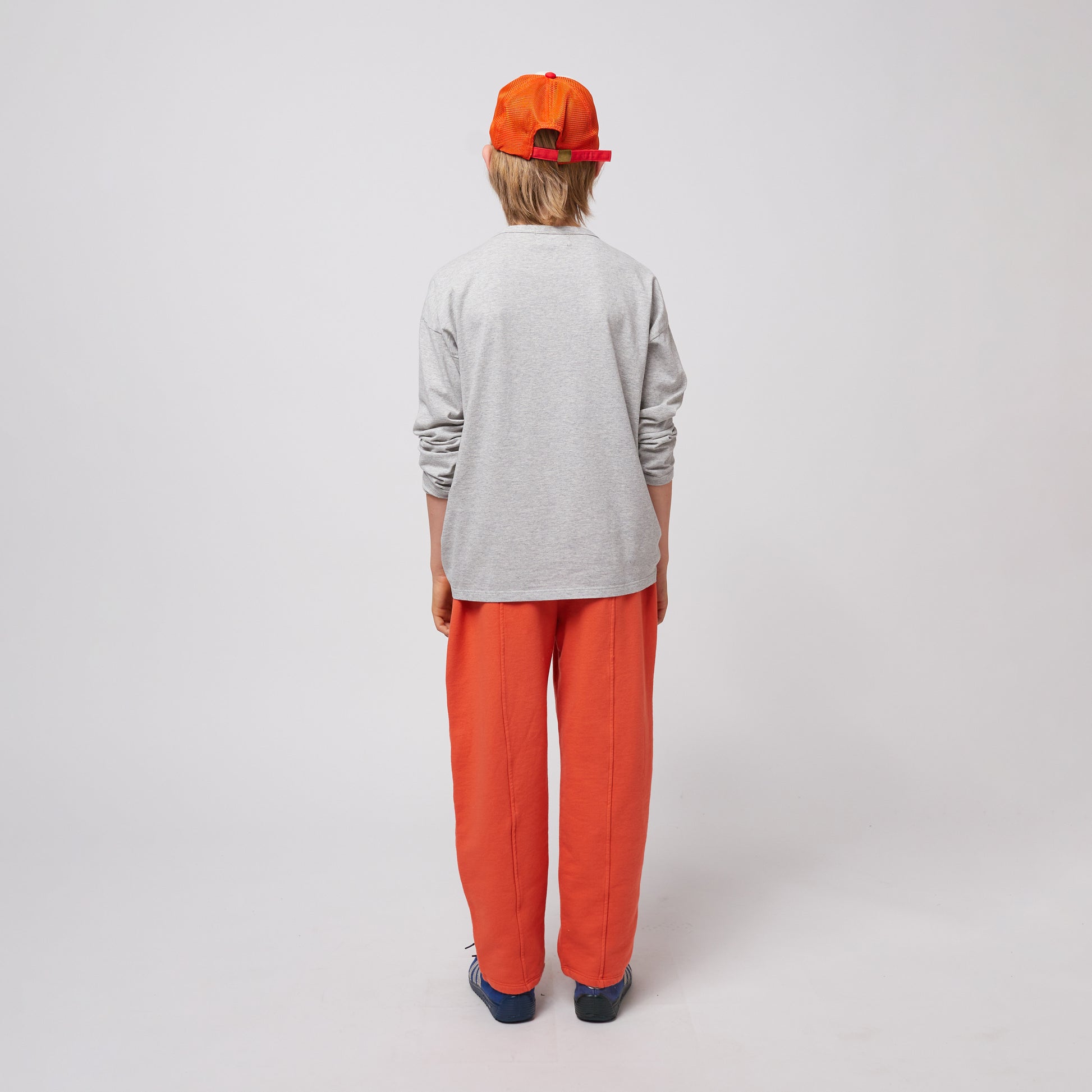 A young child is seen from the back, wearing bright orange sweatpants with stitched seams and a relaxed fit, paired with a plain grey sweatshirt. They are also wearing a red and orange trucker cap with a mesh back. Their arms hang loosely at their sides, and they wear blue Adidas sneakers with white stripes. The background is plain white.