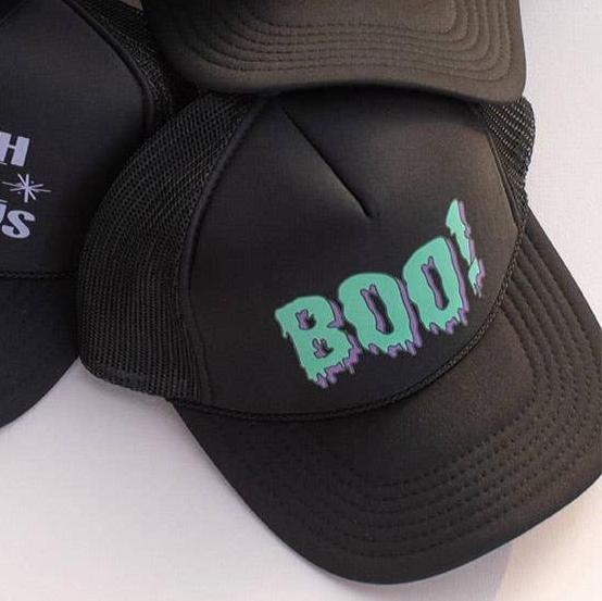 A black trucker cap with snap fastening and mesh back featuring green spooky style writing of th word BOO! surrounded by purple so it looks like dripping slime