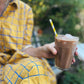 Person holding a glass of chocolate milkshake with a straw outdoors