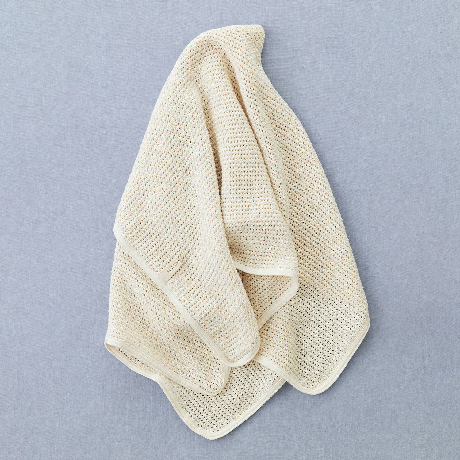 on a grey blue backdrop, a cotton linen waffle knitted blanket in a cream colour, draped in a casual way showing the folds of the material