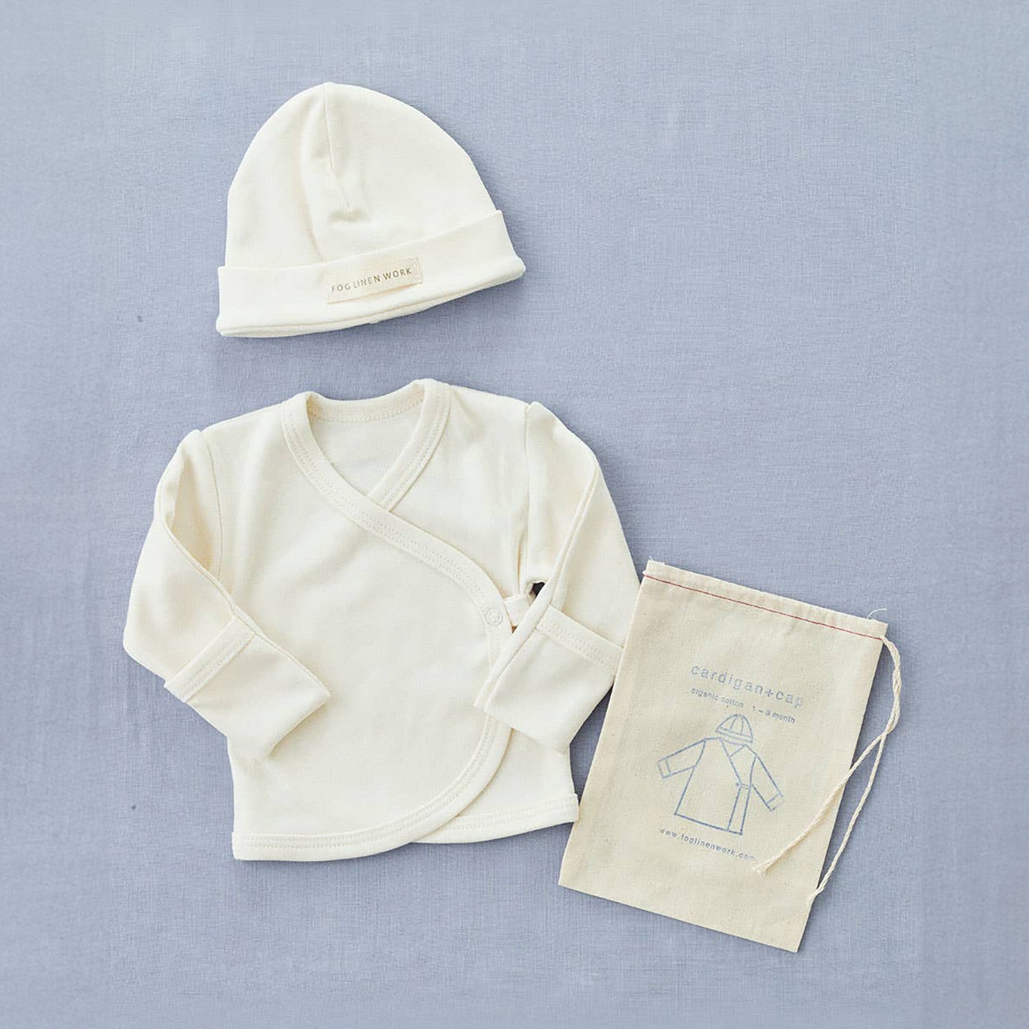 fog linen cotton baby cap and wrap front cardigan in a creamy white colour. the arms have a little hand fold over detail so babys do not scratch their skin. next to it is its draw string bag with a line illustration of the hat and cardigan and the fog linen logo